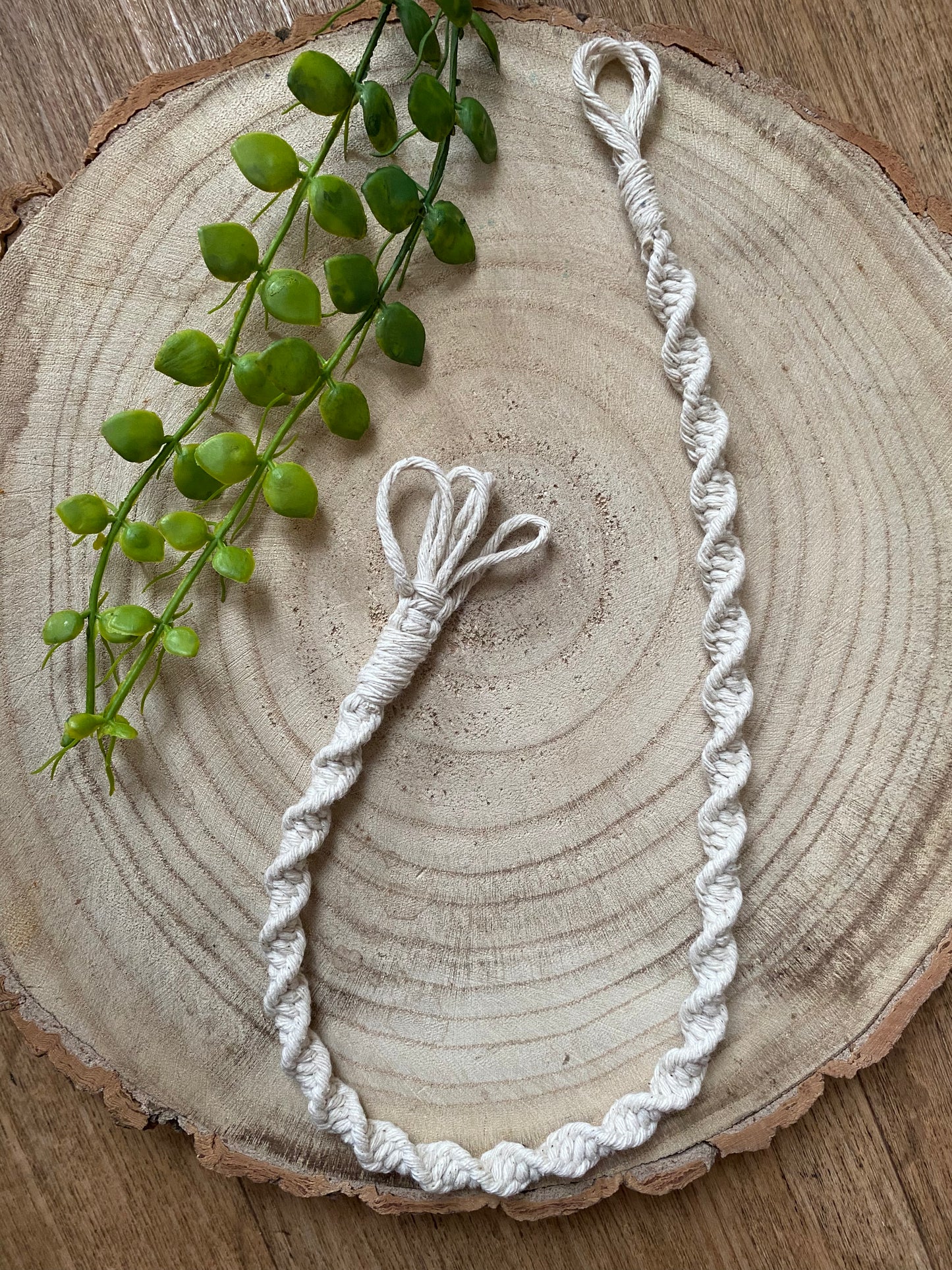 A macrame plant hanger extension made with natural cotton yarn, is laid out on a wooden surface with some green plant vines.