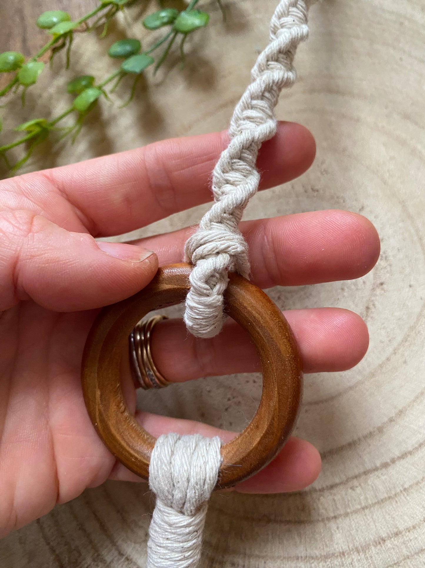 A hand holding a macrame plant hanger extension made with natural cotton yarn, which is attached to the wooden ring of a macrame plant hanger.