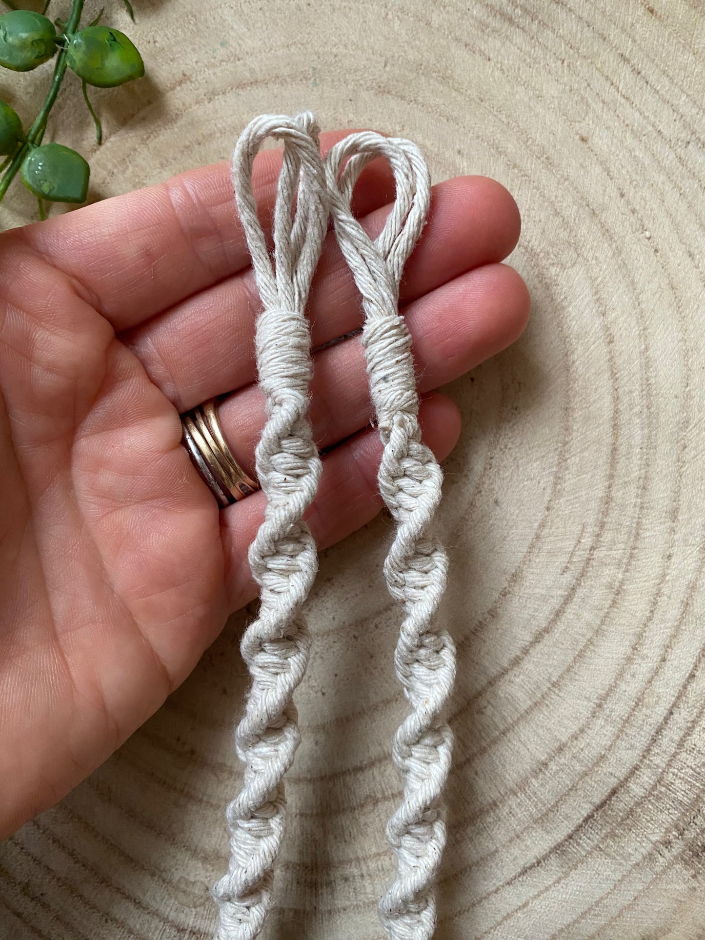 A hand holding a macrame plant hanger extension made with natural cotton yarn, on a wooden surface.