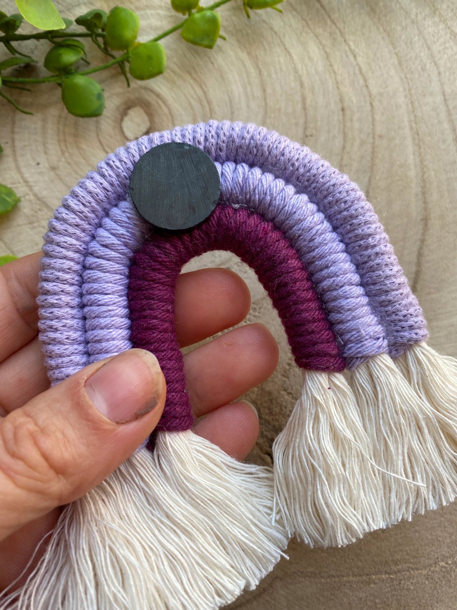 A hand holding a small handmade macrame rainbow fridge magnet in soft cotton purple cords, showing the magnet on the reverse side of the rainbow.