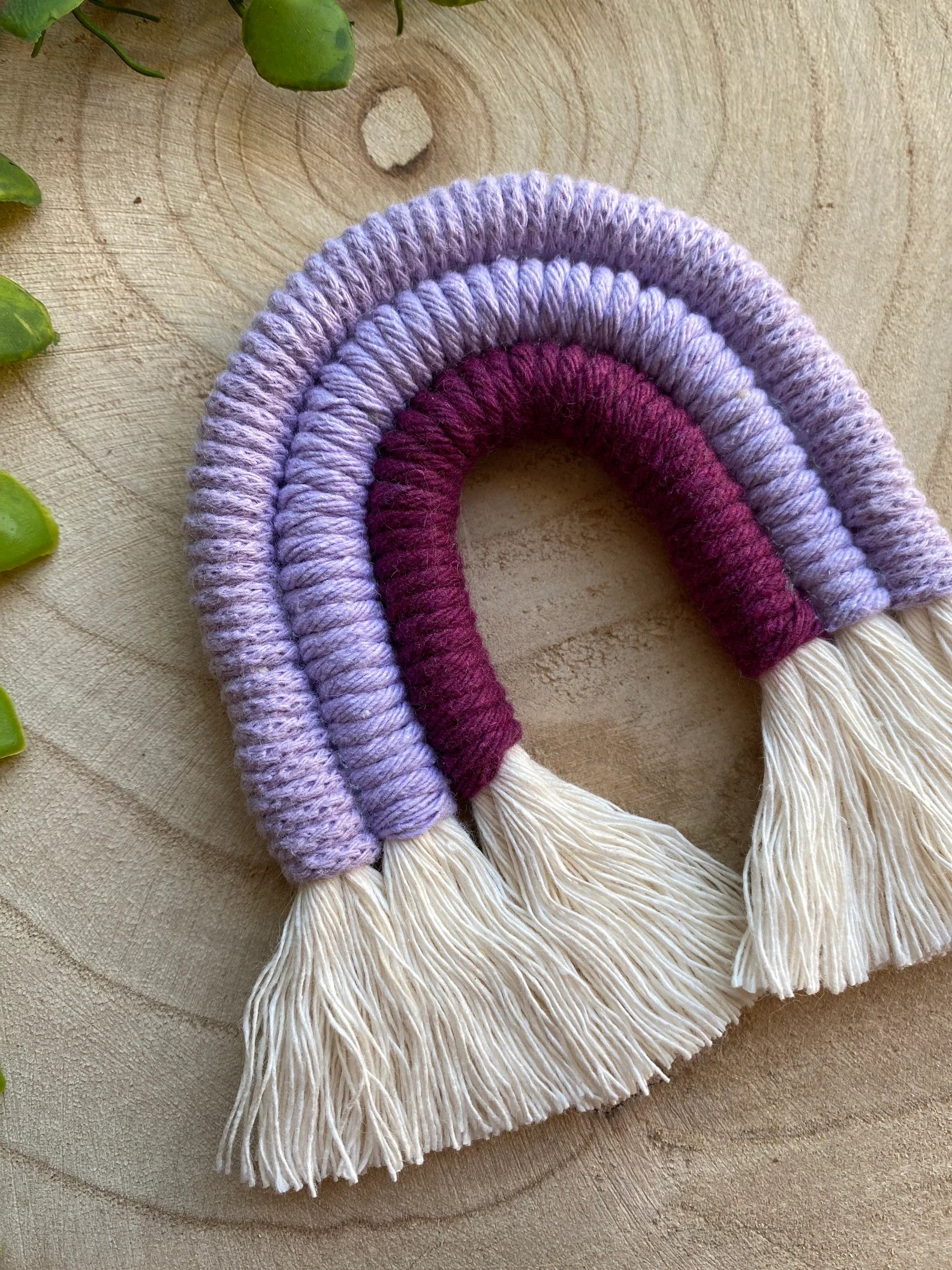 Angled close up of a small handmade macrame rainbow fridge magnet in soft cotton purple cords, on a wooden surface with some green plant vines.