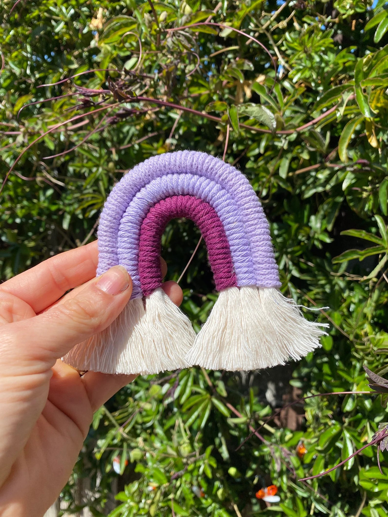 A hand holding a small handmade macrame rainbow fridge magnet in soft cotton purple cords, in front of lush green plants outside.