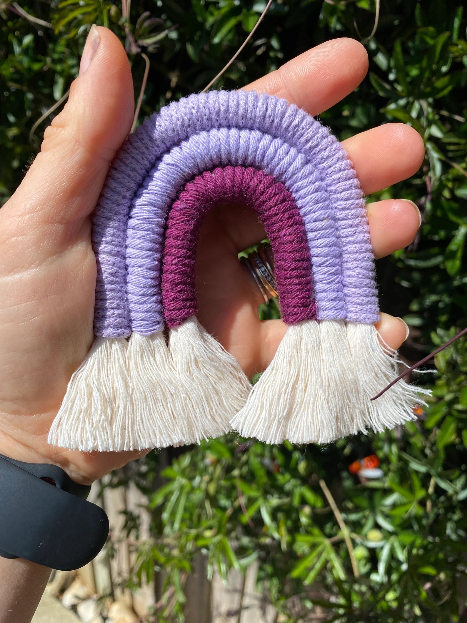 A hand holding a small handmade macrame rainbow fridge magnet in soft cotton purple cords, outside in the sunshine.