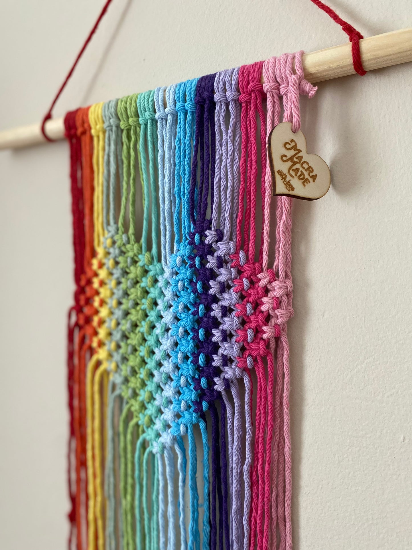 A close up image of a macrame wall hanging with heart detail, made with rainbow colours of yarn on a wooden dowel, hanging on a beige wall.