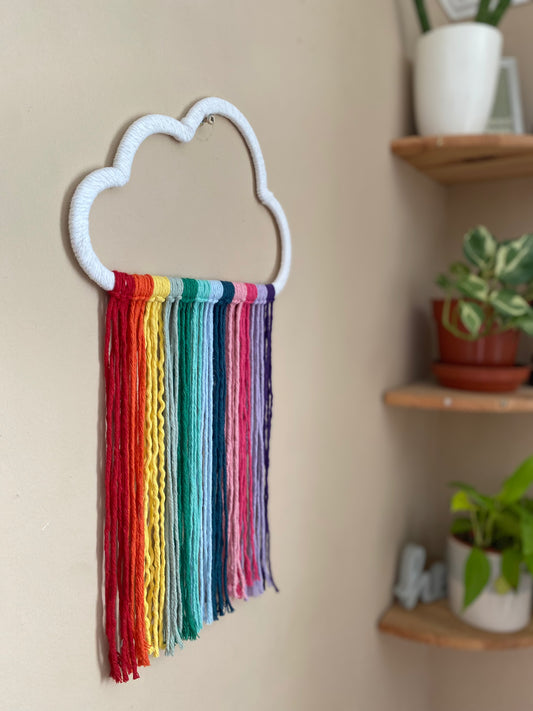 A cloud shaped wall hanging decoration, made with rainbow hanging tassels, displayed against a beige wall.