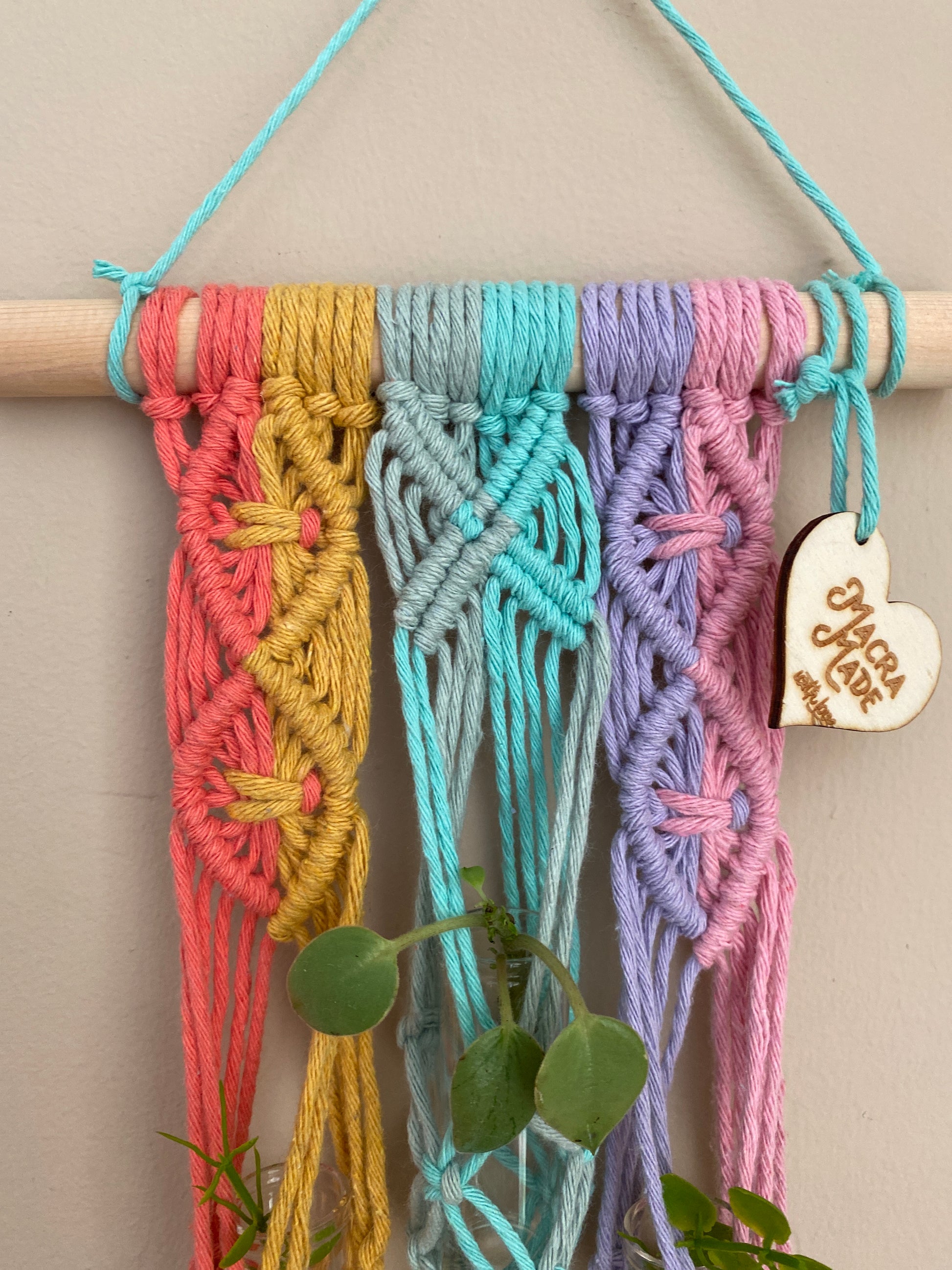 Close up of the intricate knots on a handmade macrame wall hanger houseplant propagation station made with pastel coloured yarn, against a beige wall.
