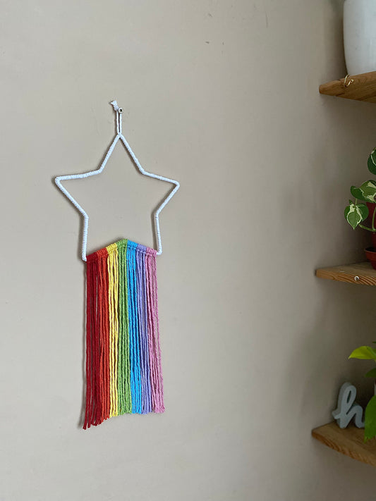 A white star frame with rainbow hanging tassels, hangs against a beige painted wall.