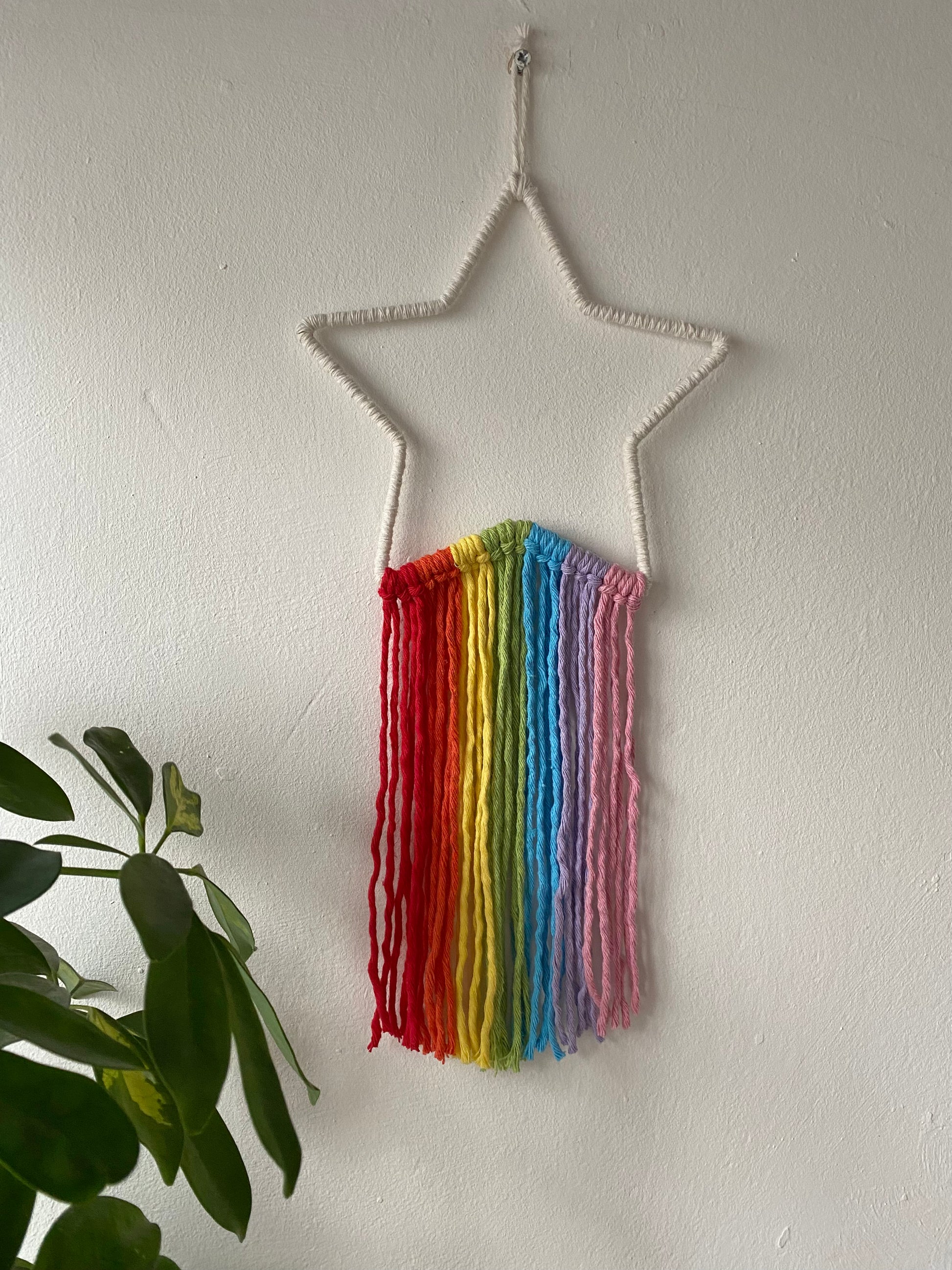 A white star frame with rainbow hanging tassels, hangs against a white painted wall.