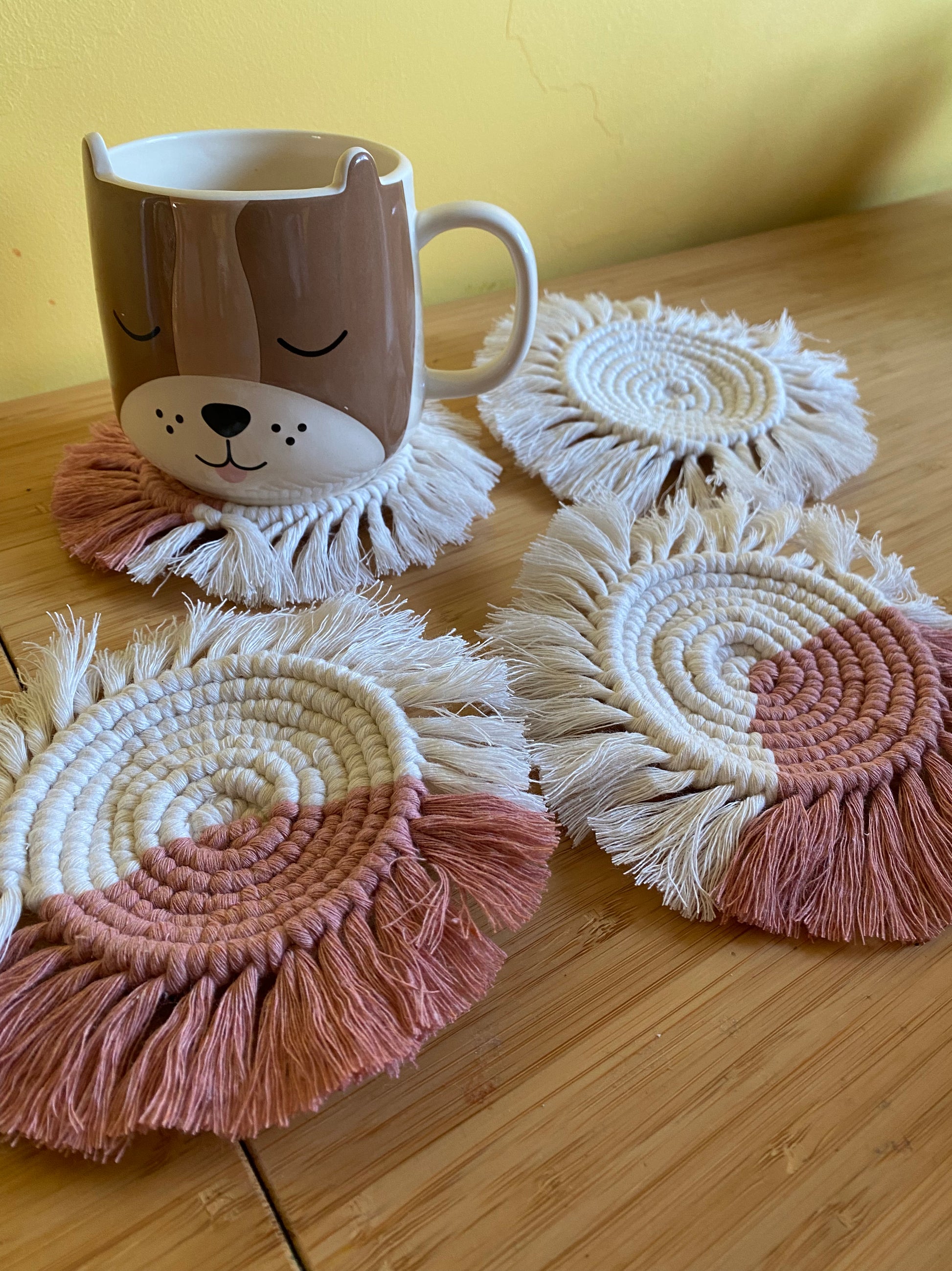 A set of 4 round macrame coasters made with blush pink and natural yarns, with a soft fringed edge, are placed on a wooden table,  with a novelty dog mug placed on top of one of the coasters.