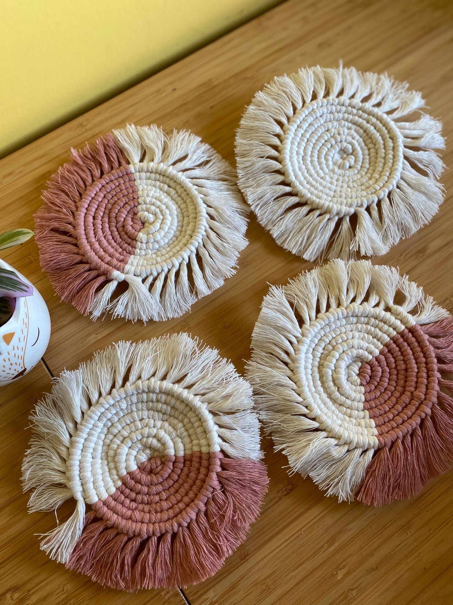 A set of 4 round macrame coasters made with blush pink and natural yarns, with a soft fringed edge, are placed on a wooden table.