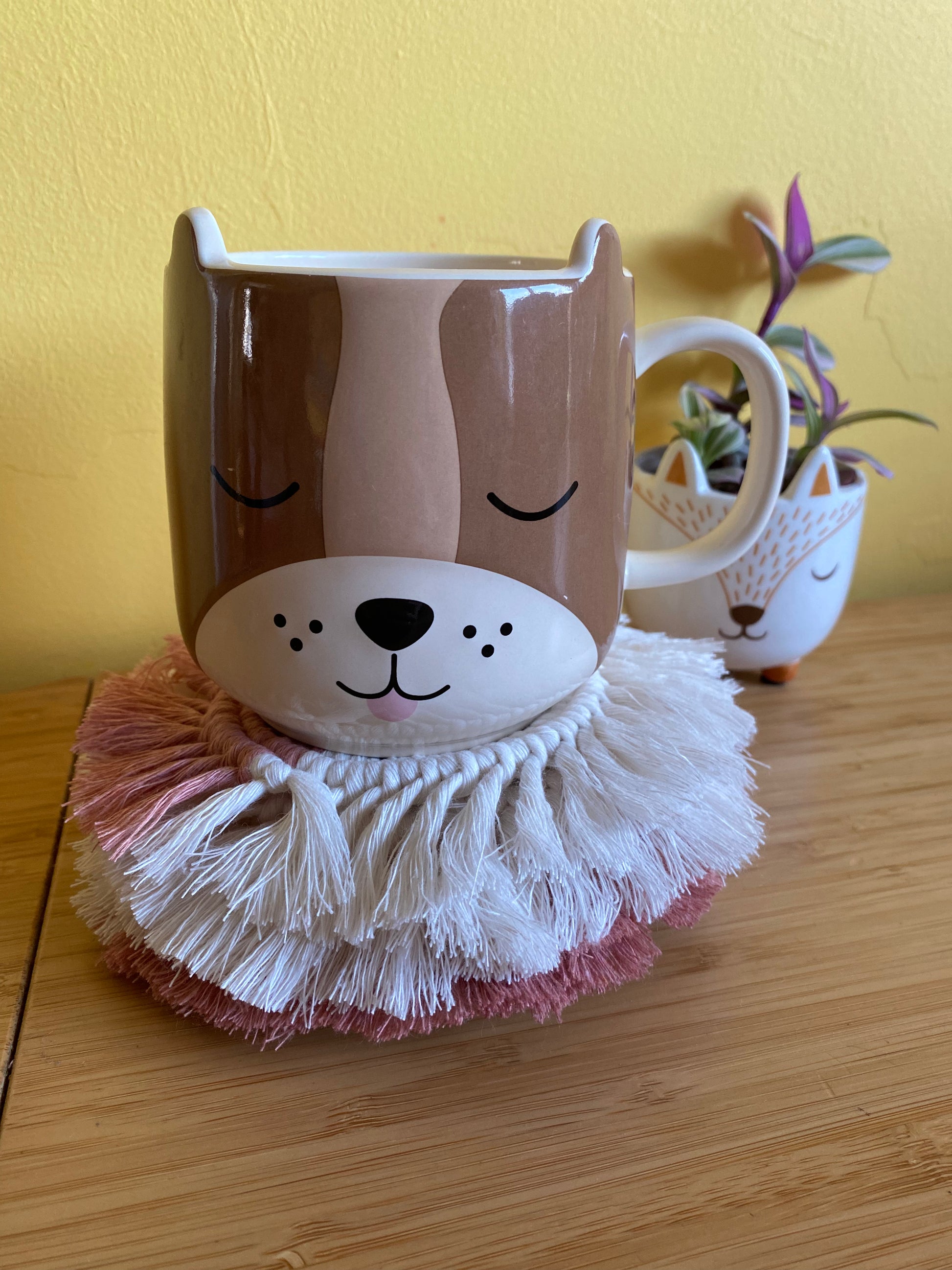 A set of 4 round macrame coasters made with blush pink and natural yarns, with a soft fringed edge, are placed on a wooden table, stacked on top of one another, with a large novelty dog mug on top of the pile. There is a small houseplant in a ceramic plant pot in the background.