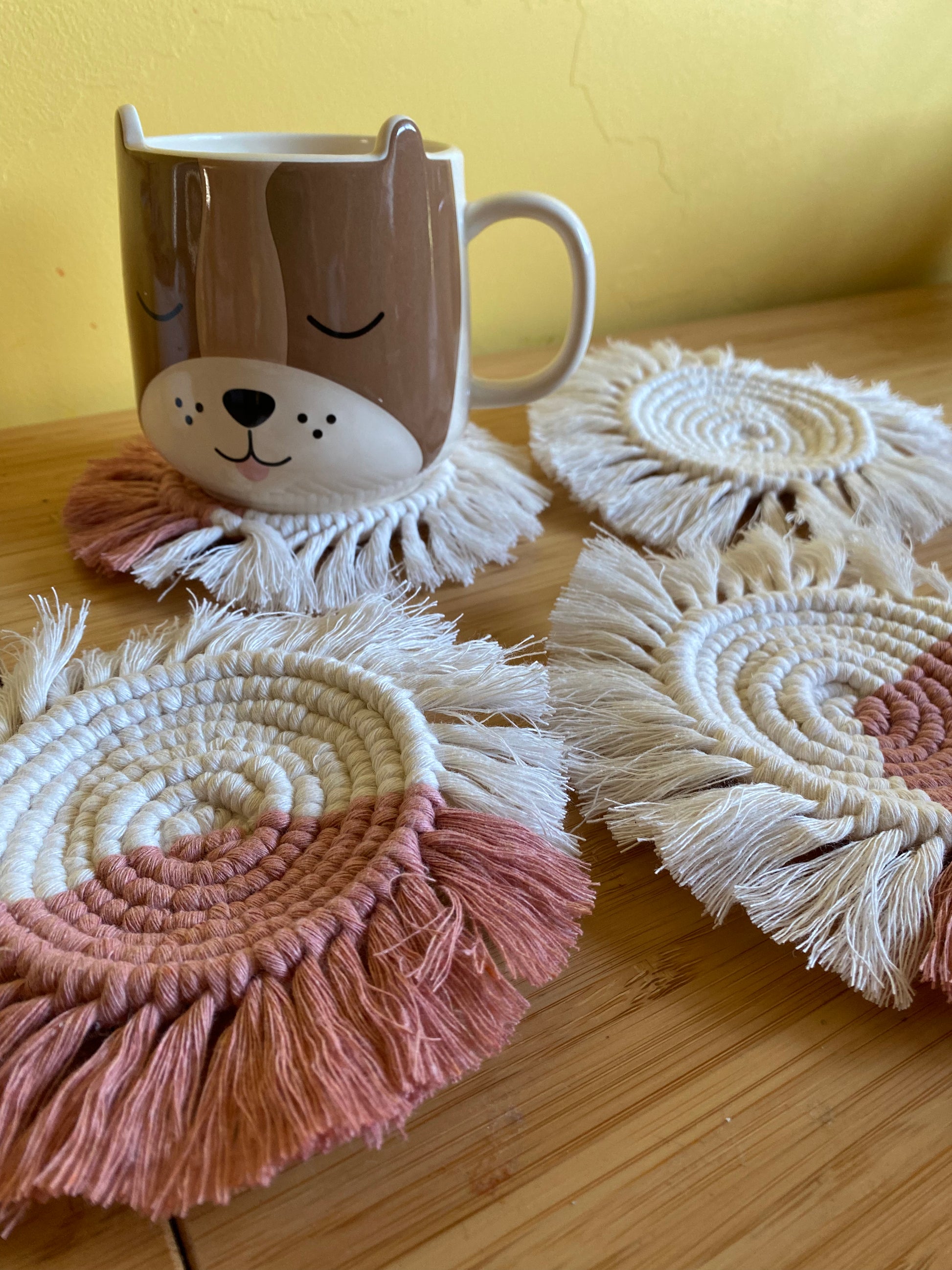 A close up image of a set of 4 round macrame coasters made with blush pink and natural yarns, with a soft fringed edge, are placed on a wooden table, with a novelty dog mug placed on top of one of the coasters.