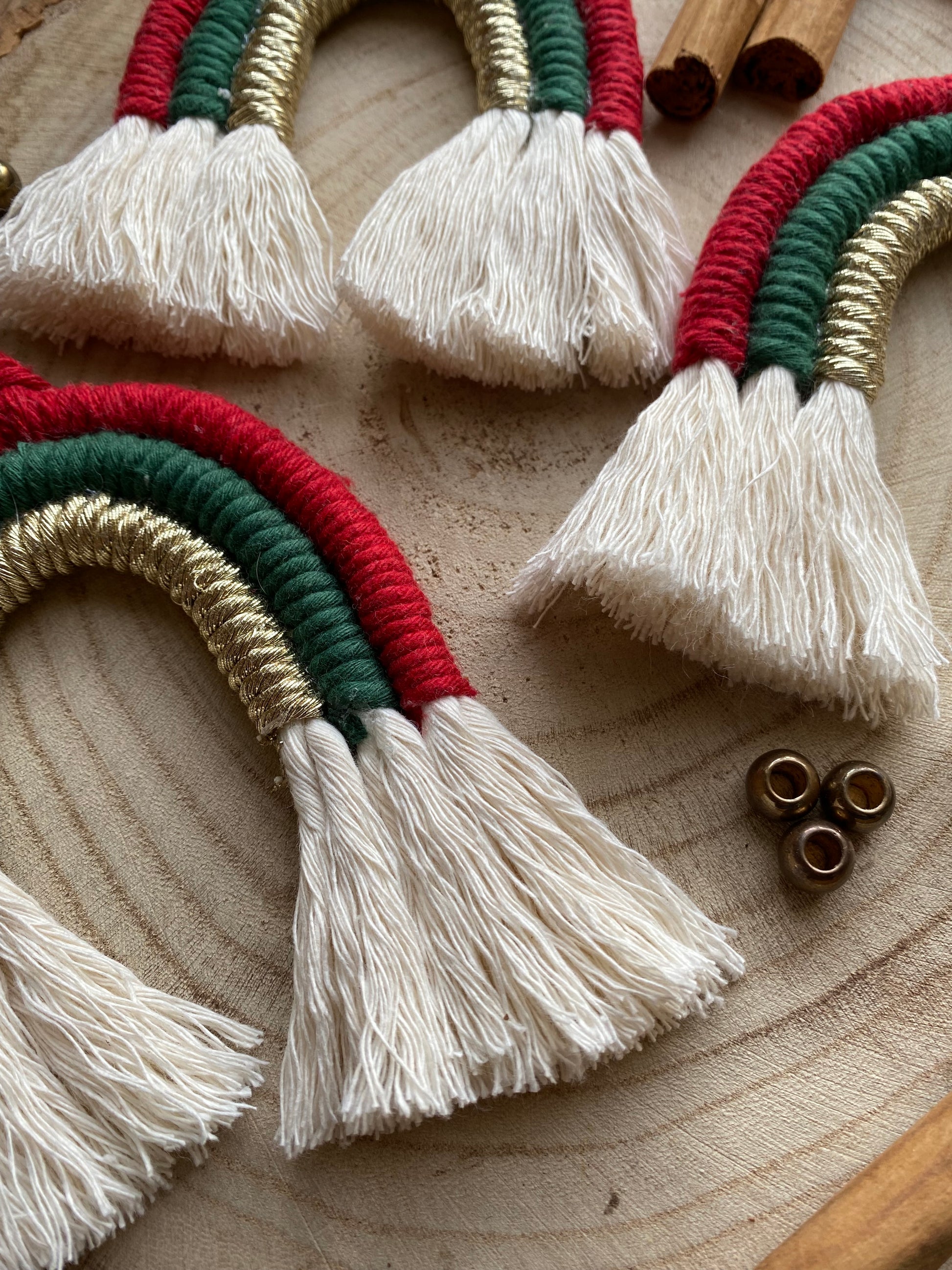 Close up of a set of three handmade macrame rainbow Christmas decorations in red, green, and gold yarn with white tassels, displayed on a wooden surface.