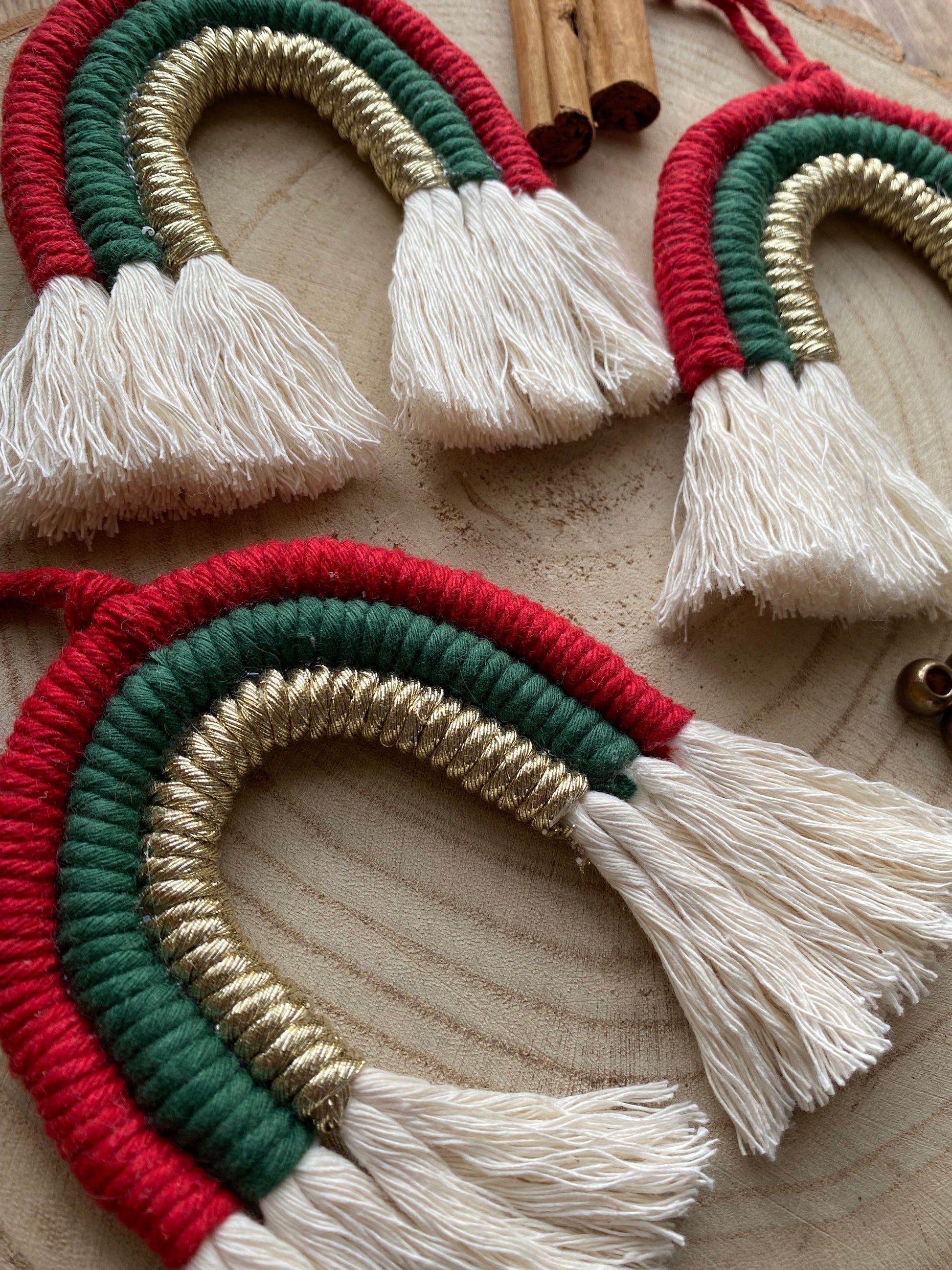 Angled close up of a set of three handmade macrame rainbow Christmas decorations in red, green, and gold yarn with white tassels, displayed on a wooden surface.