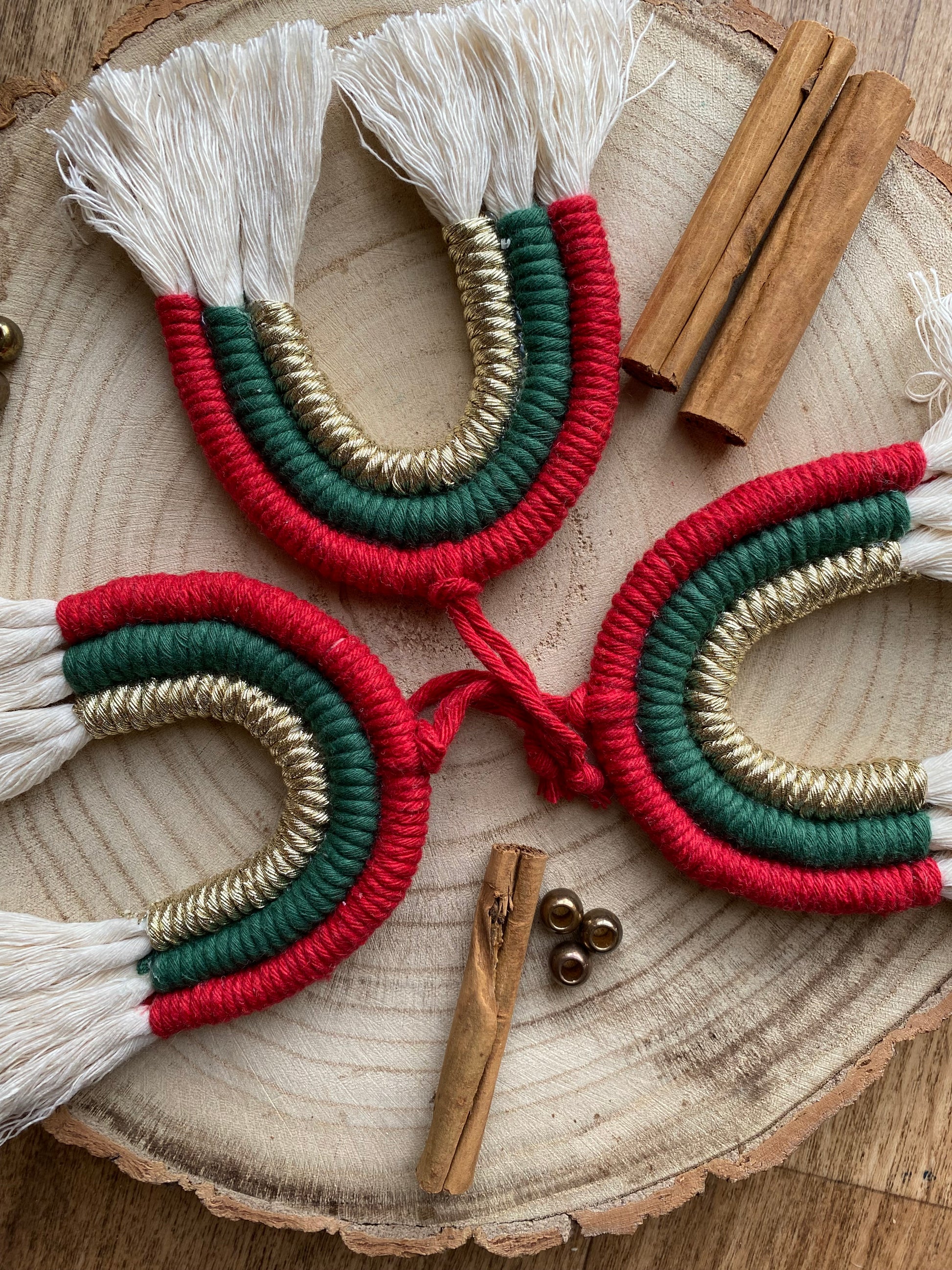 A set of three handmade macrame rainbow Christmas decorations in red, green, and gold colours with white tassels, displayed on a wooden surface.