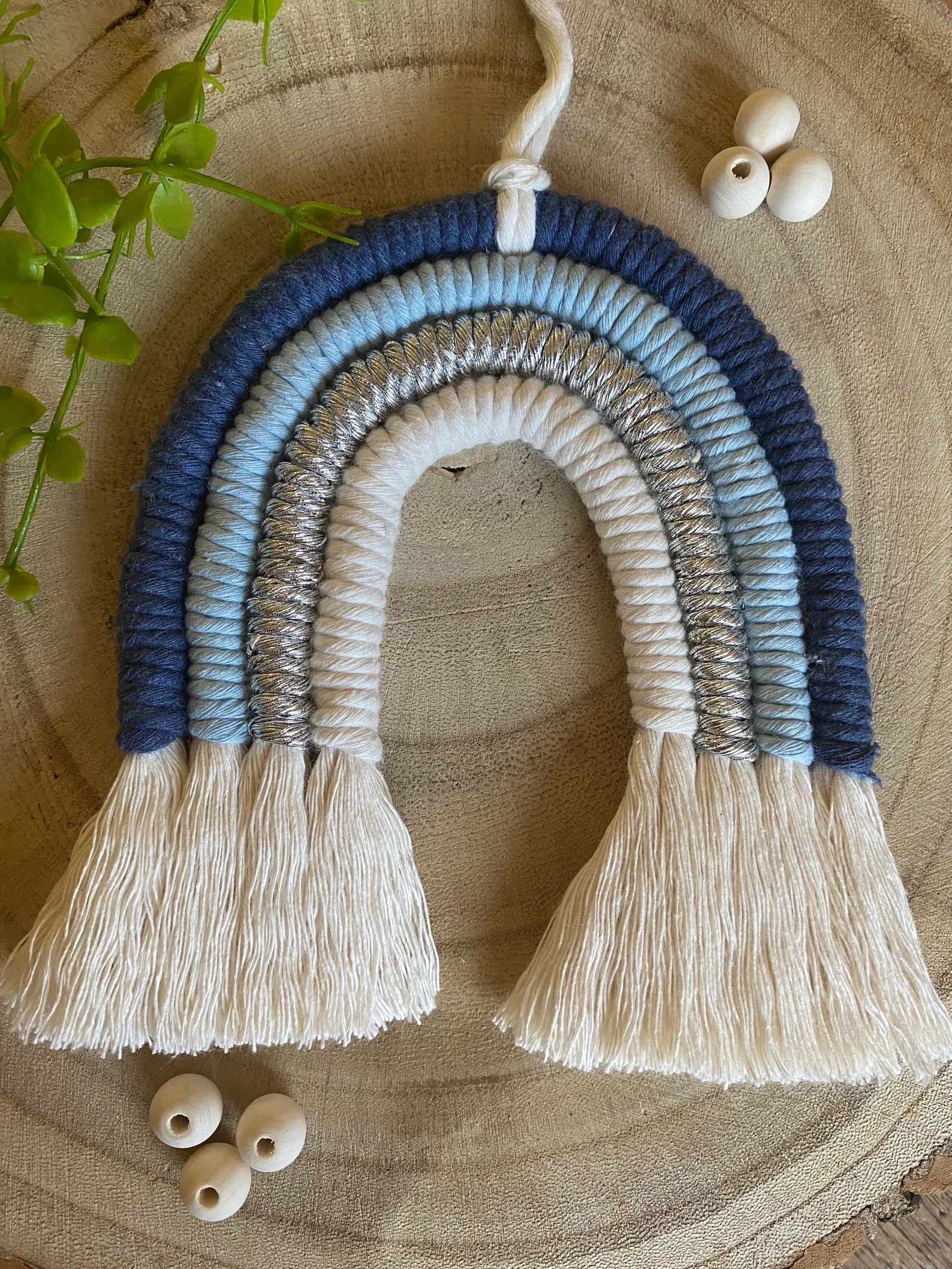 Handmade blue macramé rainbow wall hanging made with recycled cotton cord in soft blue and neutral tones, shown against a wooden background surrounded by beads and some green plant vines.