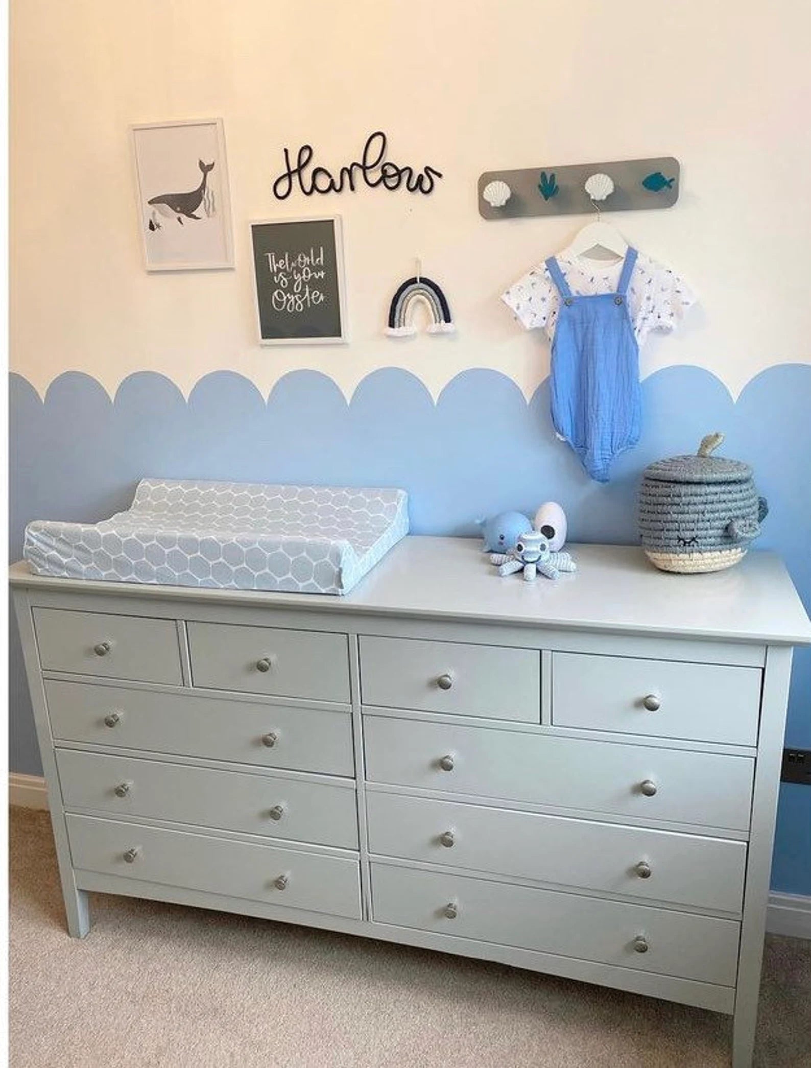 A nursery scene including large chest of drawers, with changing mat and accessories on the top. The wall behind the drawers is cream and blue, and has various prints hanging on the it, plus a little outfit on a hanger and a small blue macrame rainbow.