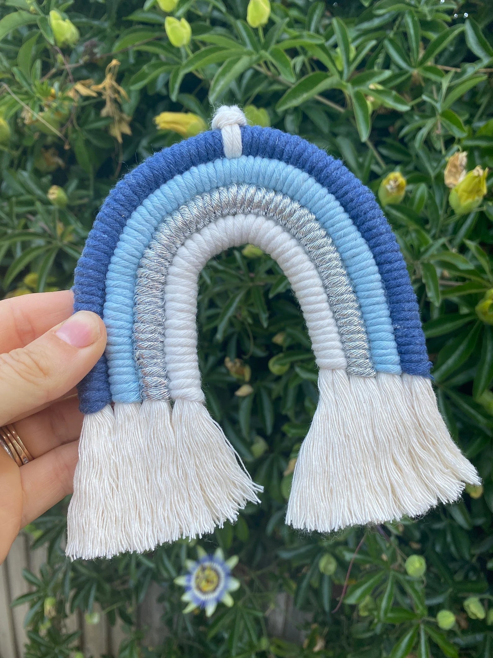 A hand holding a handmade blue macramé rainbow wall hanging made with soft cotton cord against a green plant background.