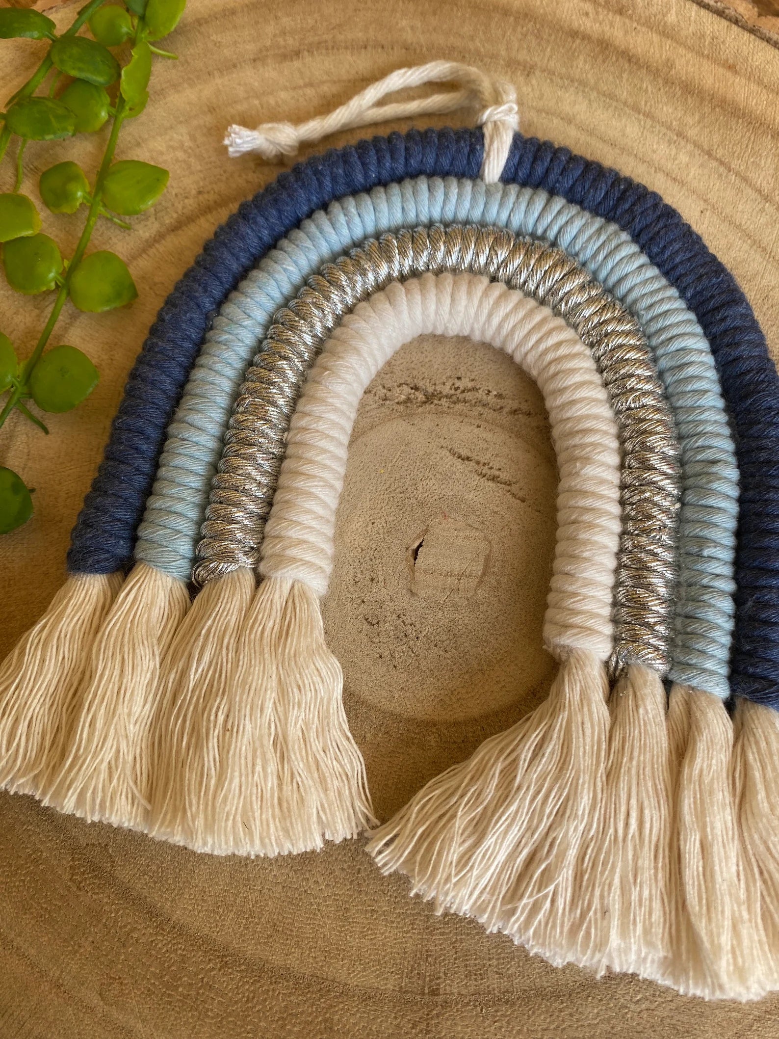 Handmade blue macramé rainbow wall hanging made with cotton cord in soft blue and neutral tones, shown against a wooden background.