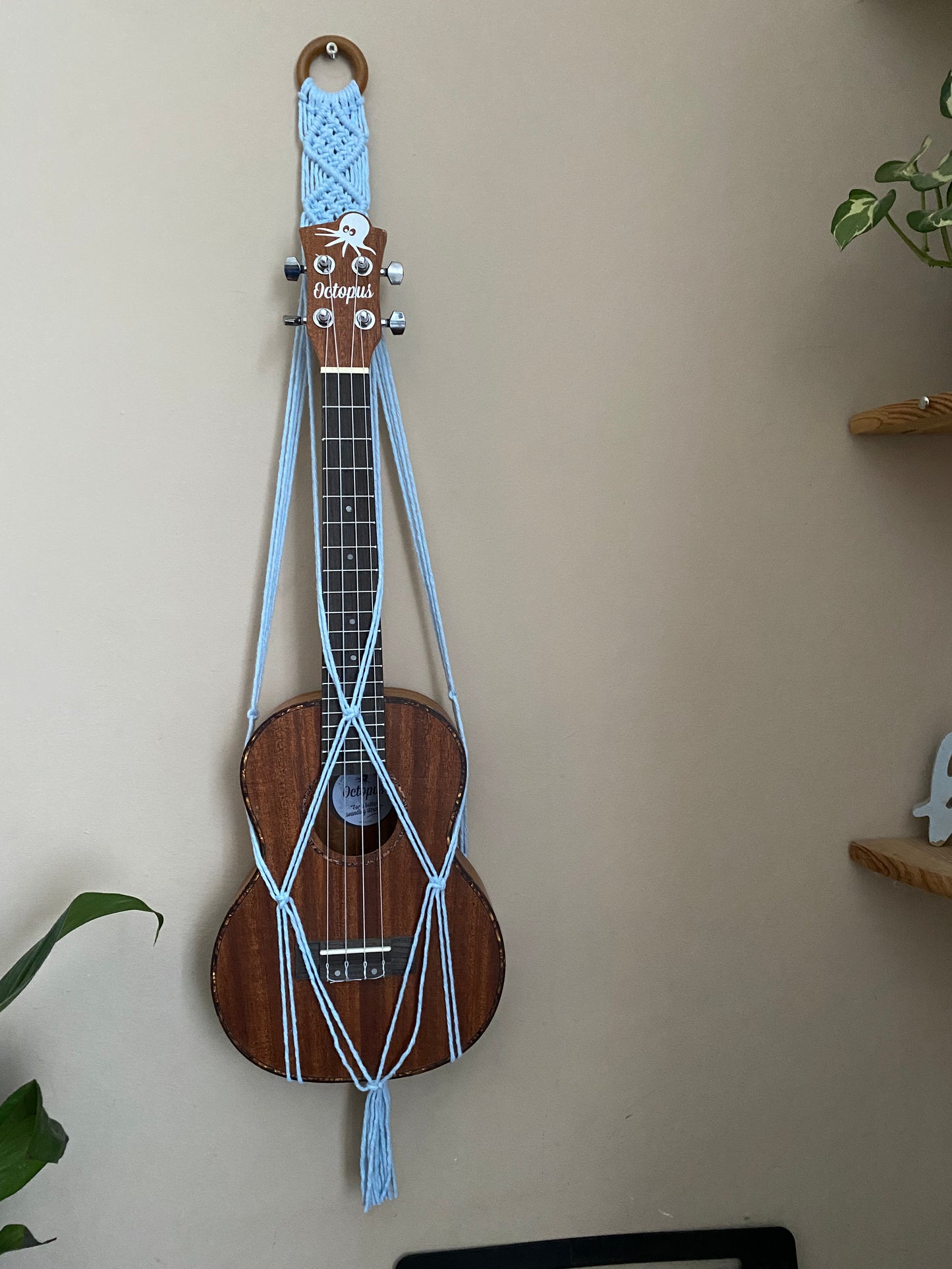 A dark brown ukulele sits inside of a handmade macrame wall hanging ukulele hanger, which is made with light blue recycled cotton, and a repurposed wooden hanging ring.