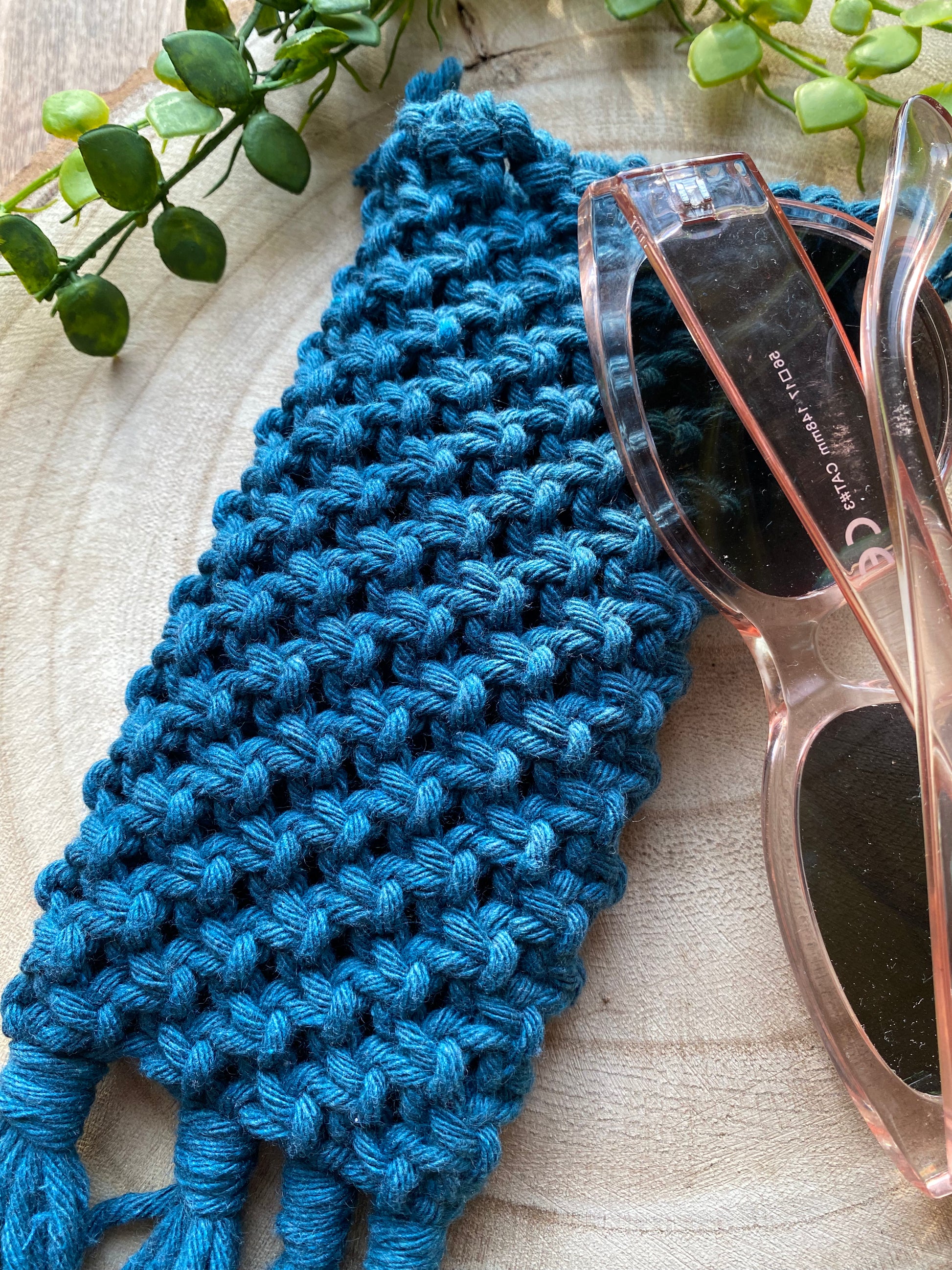 Handmade macrame soft glasses pouch made with petrol blue cotton yarn. Displayed with a pair of pink sunglasses laid across it, on a round wooden surface.