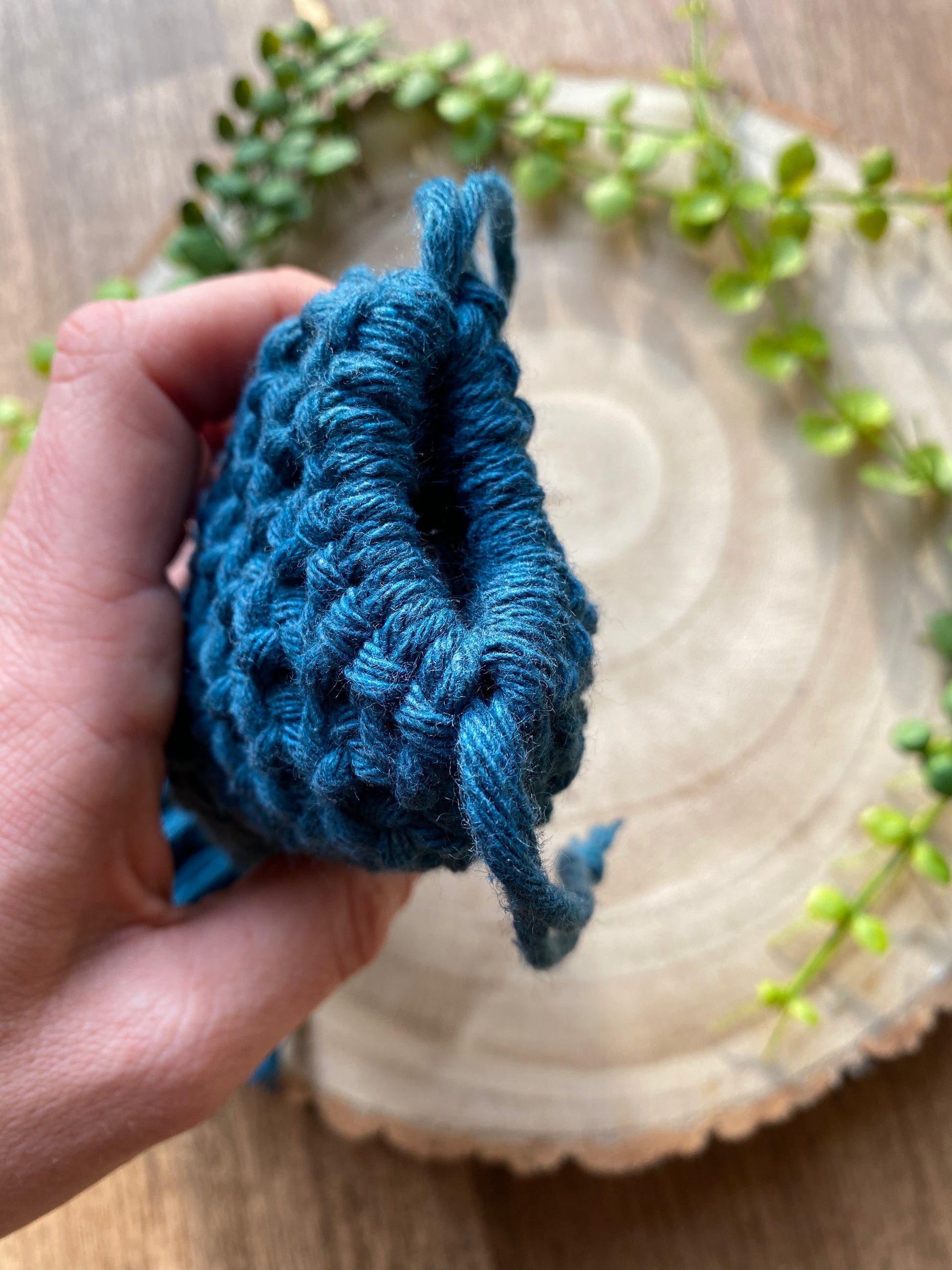 A hand holding a macrame glasses pouch made with petrol blue recycled cotton. The image focuses on the top drawstring opening of the glasses pouch.