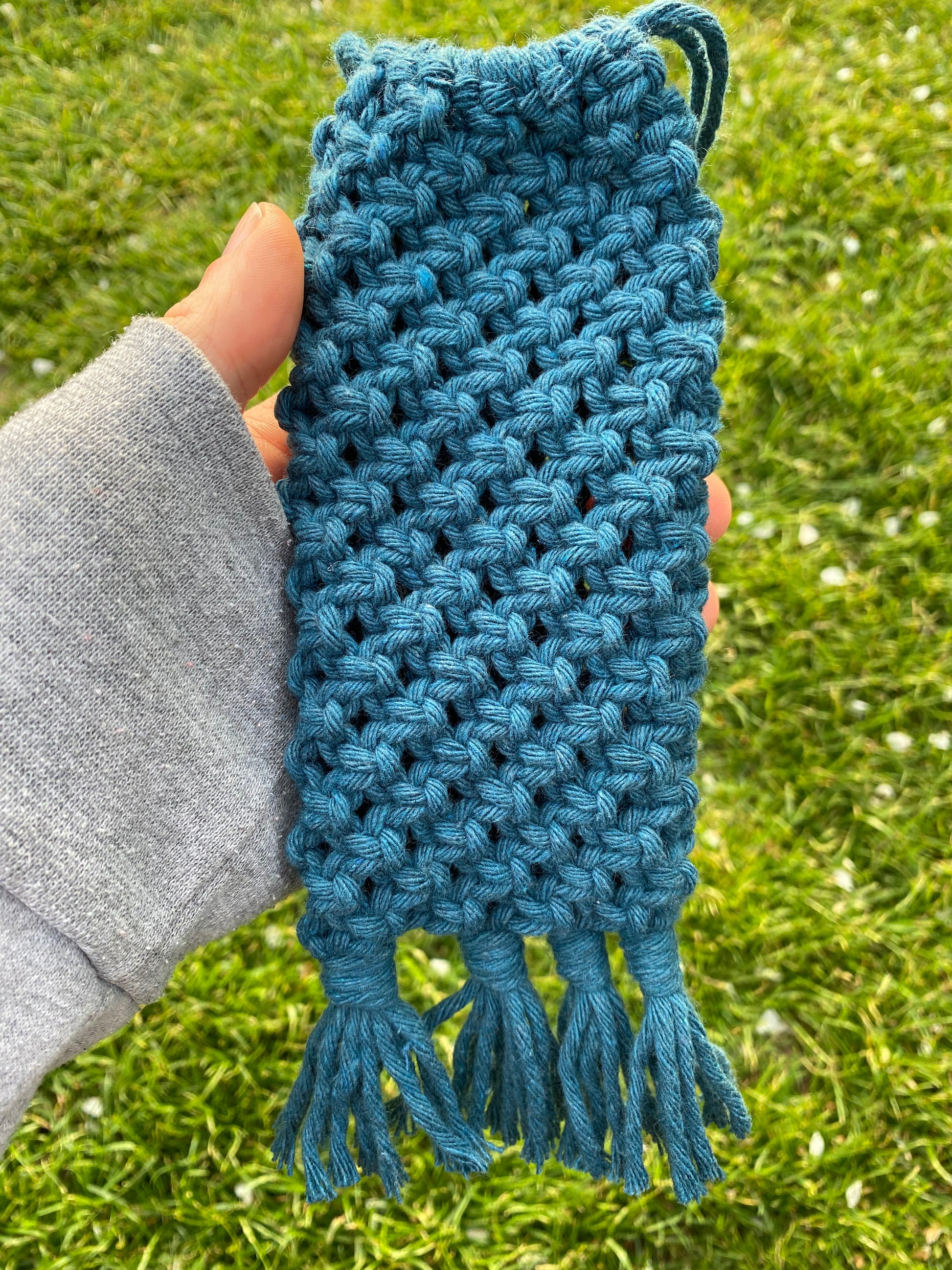 A hand holding a handmade macrame soft glasses pouch made with petrol blue cotton yarn, outside with lush green grass in the background.