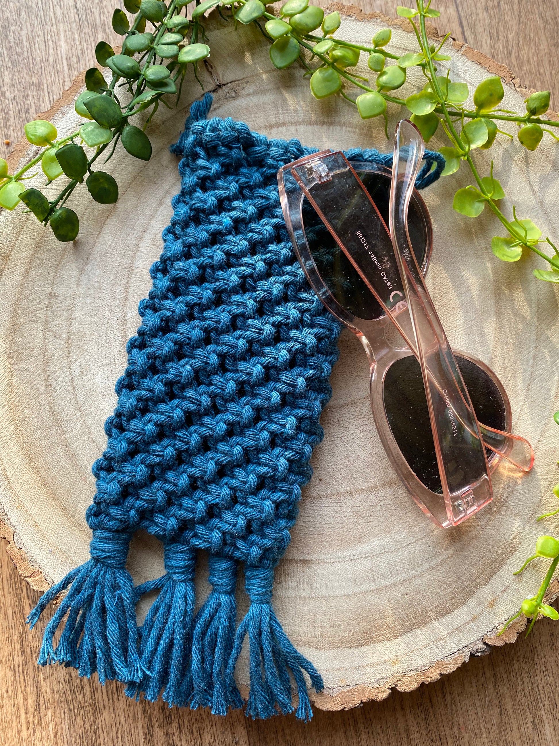Handmade macrame soft glasses pouch made with petrol blue cotton yarn. Displayed with a pair of pink sunglasses beside it, on a round wooden surface.
