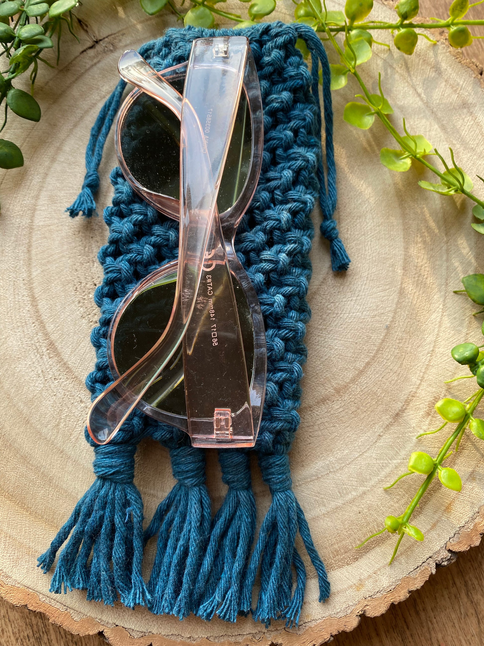 Handmade macrame soft glasses pouch made with petrol blue cotton yarn. Displayed with a pair of pink sunglasses on top of it on a round wooden surface.