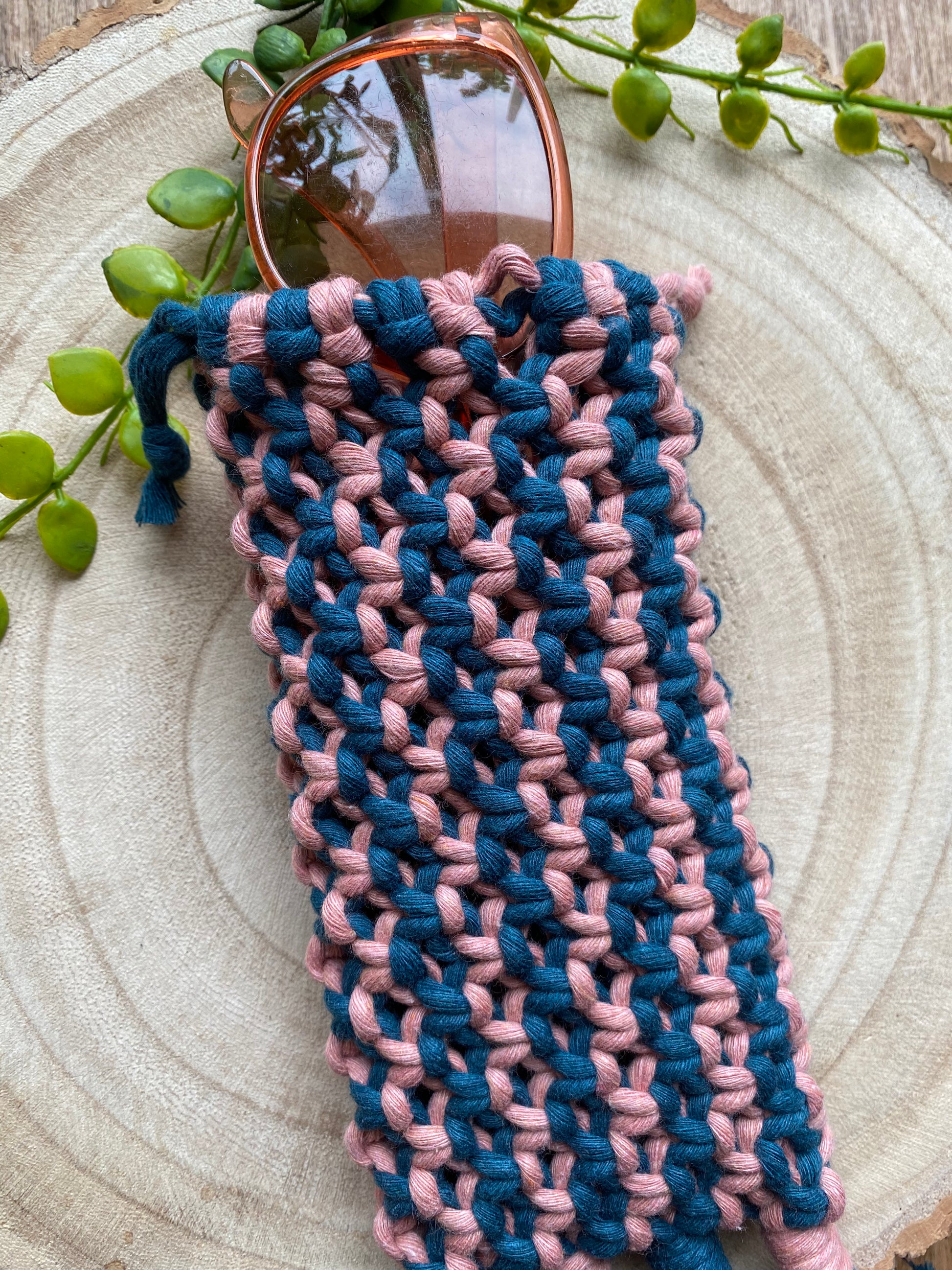 Handmade macrame soft glasses pouch made with pink and blue cotton yarn. Displayed with a pair of pink sunglasses just showing out of the top of the pouch, on a round wooden surface.