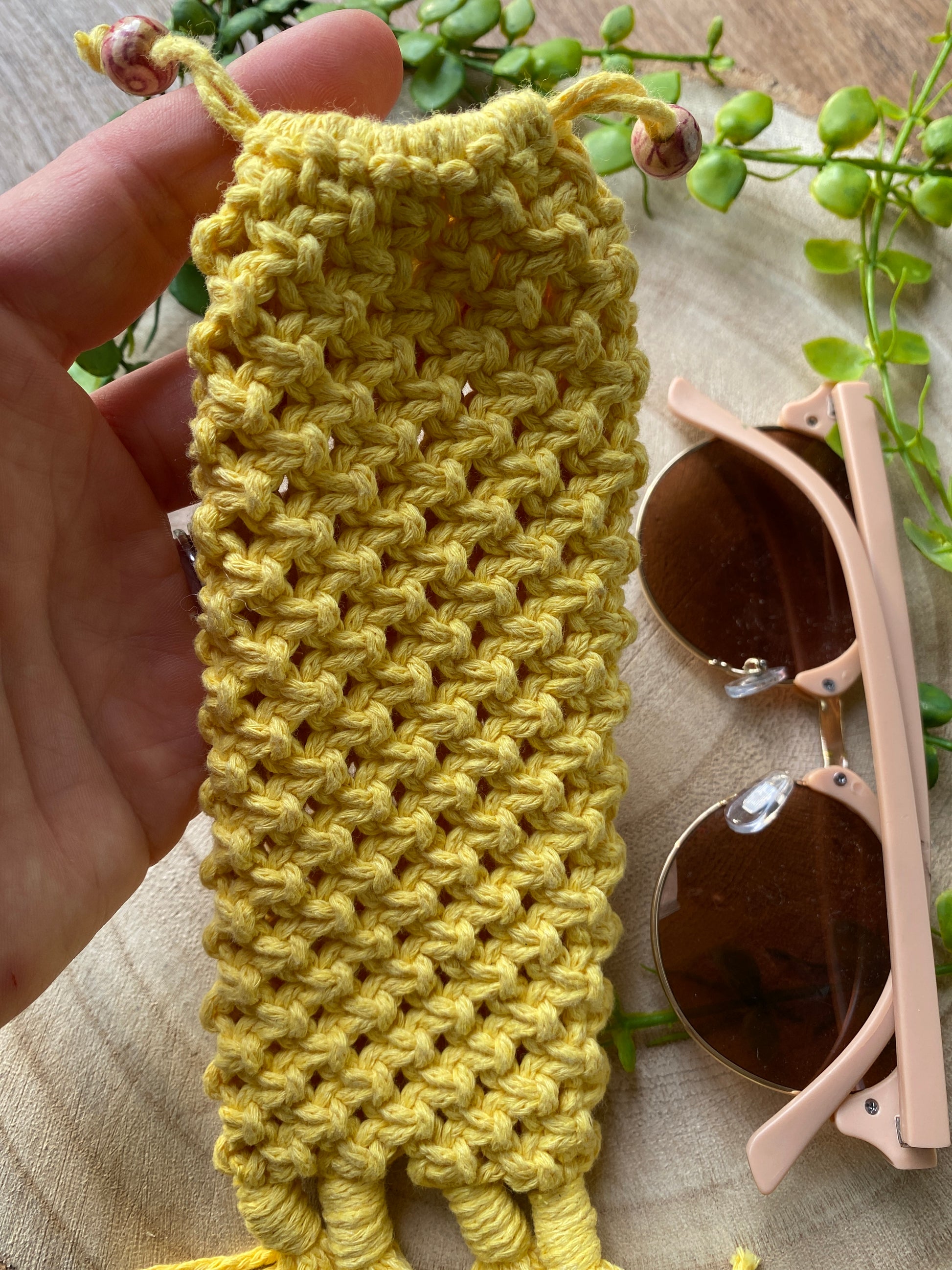 A hand holding a handmade macrame soft glasses pouch made with yellow cotton yarn. Displayed with a pair of pink sunglasses laid next to it on a round wooden surface.
