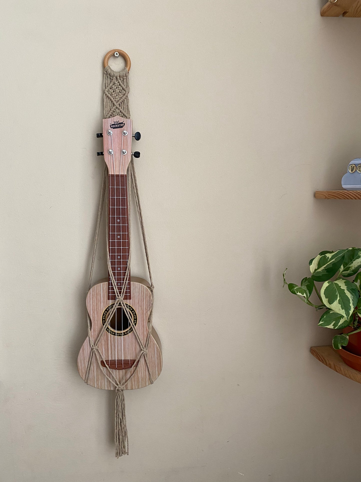 A light brown ukulele sits inside of a handmade macrame wall hanging ukulele hanger, which is made with taupe recycled cotton, and a repurposed wooden hanging ring. A small green leafy plant sits on a shelf nearby.