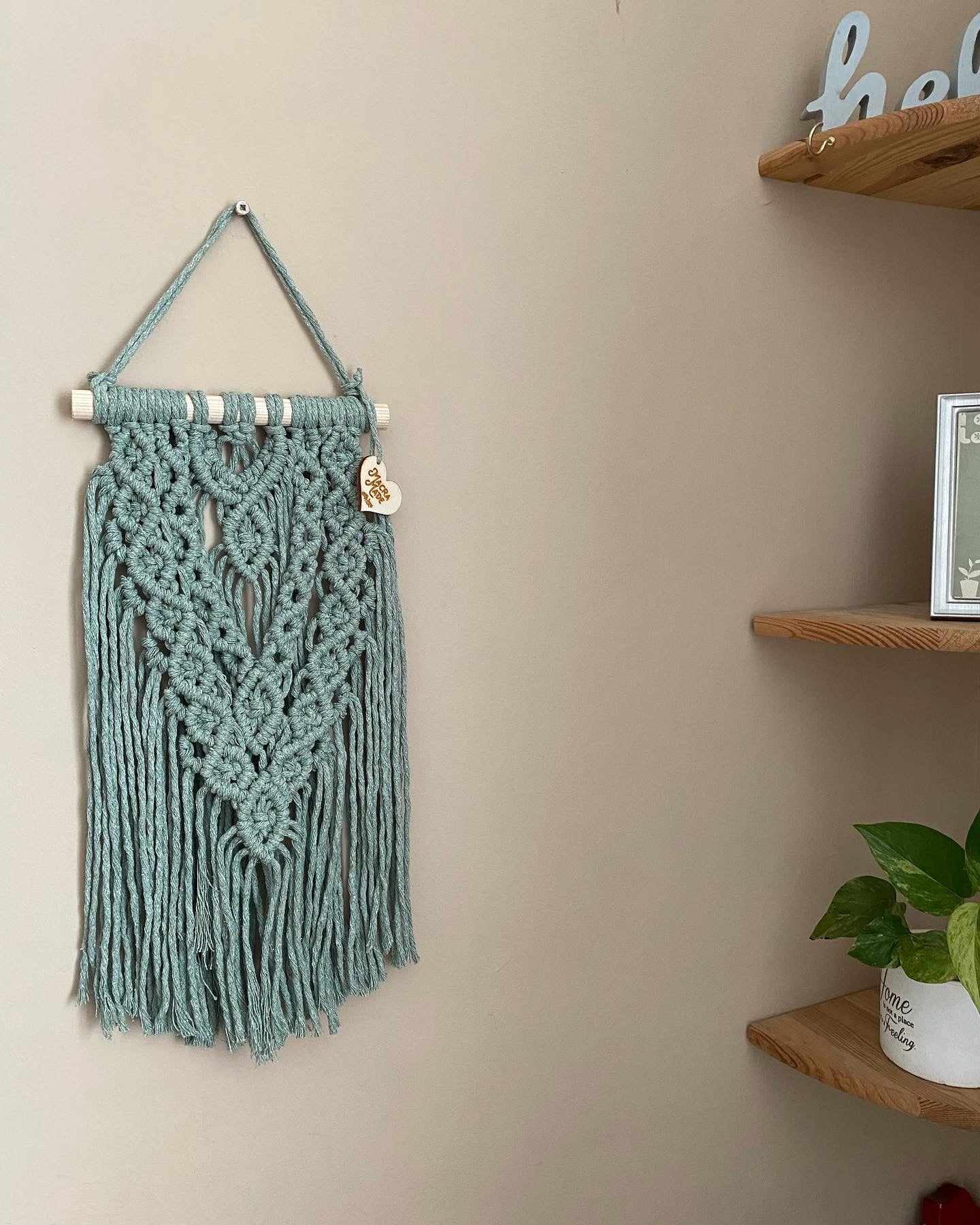 A textured, layered macrame wall hanging made with intricate knot patterns using pastel green yarn on a wooden dowel, hanging against a beige wall.