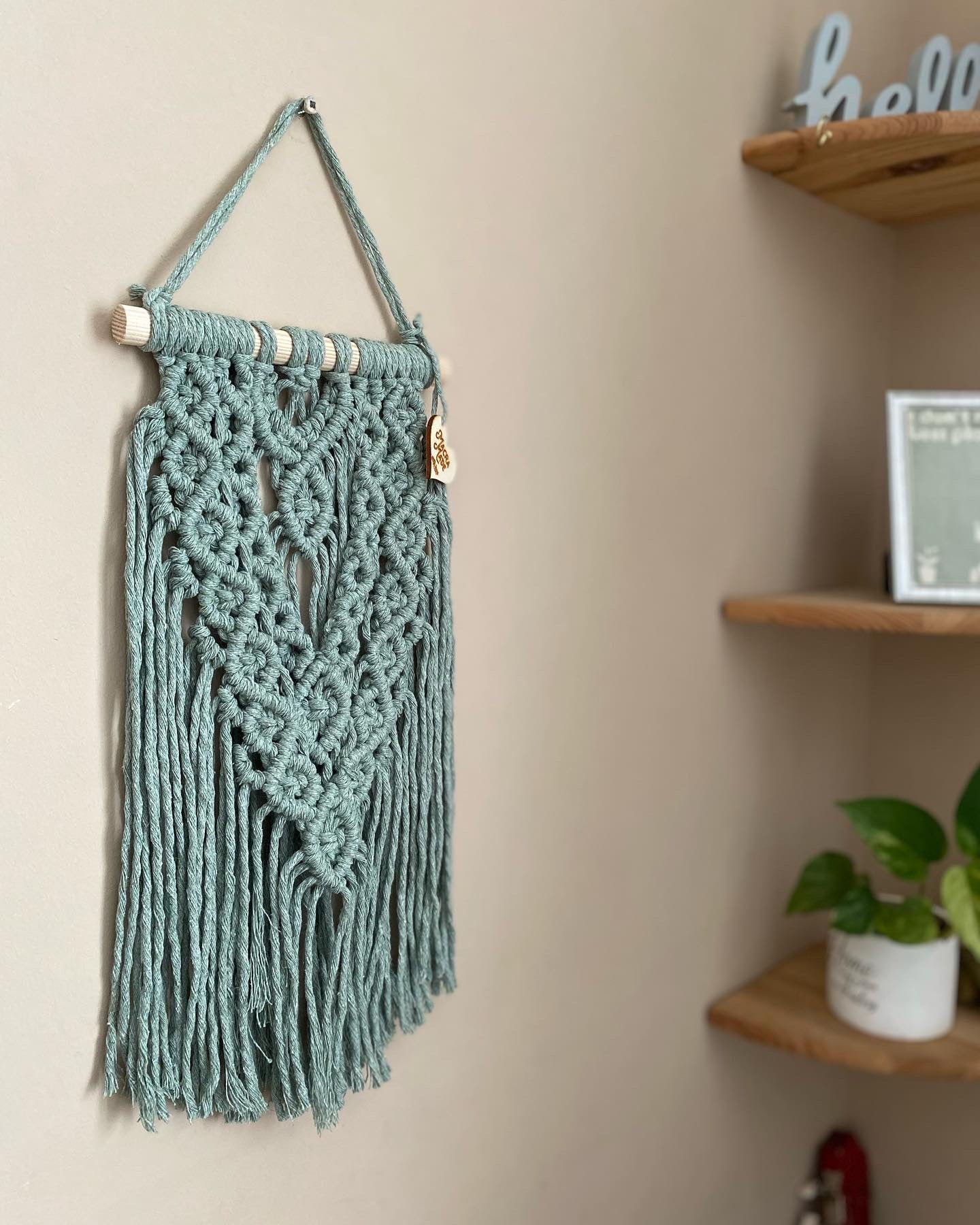 A textured, layered macrame wall hanging made with intricate knot patterns using pastel green yarn on a wooden dowel, hanging against a beige wall next to a set of corner shelves.