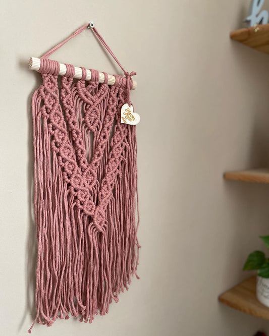 A textured, layered macrame wall hanging made with intricate knot patterns using blush pink yarn on a wooden dowel, hanging against a beige wall.