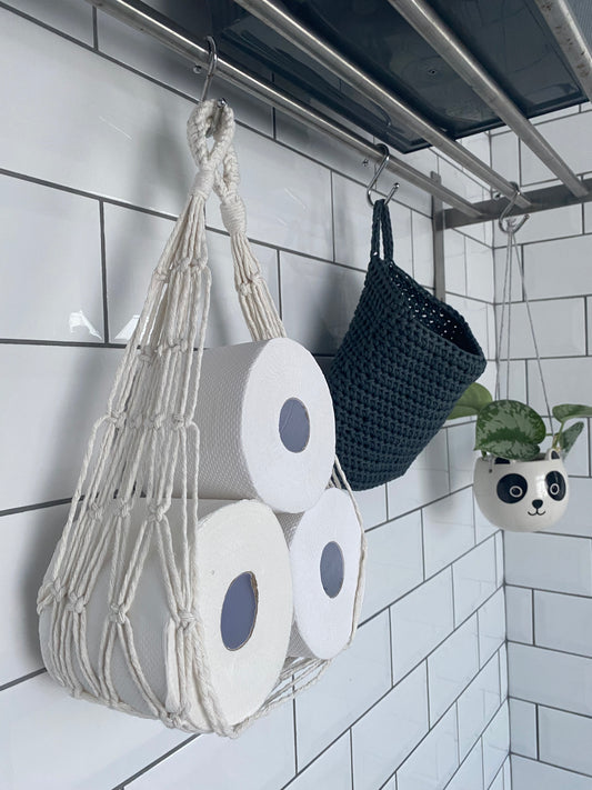 A handmade macrame toilet roll hammock style holder, made with natural coloured recycled cotton, holding three white toilet rolls. The hammock is hung from a metal hook under a metal shelf, against a white tiled background.
