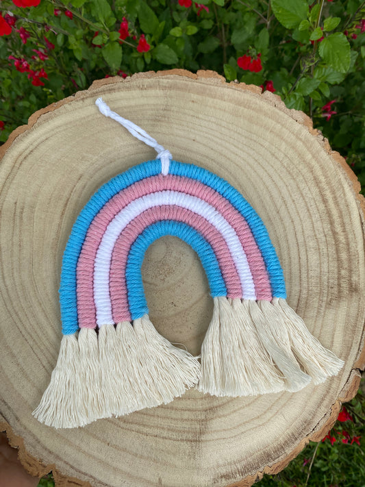 Handmade macrame rainbow made with 5 arches of recycled cotton in blue, pink and white yarns to represent the colours of the trans pride flag, displayed on a wooden board.