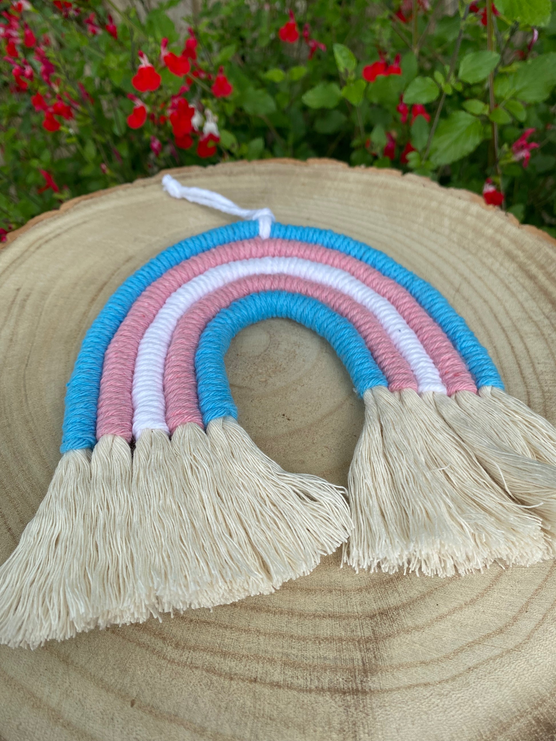 Handmade macrame rainbow made with 5 arches of recycled cotton in blue, pink and white yarns to represent the colours of the trans pride flag, with combed out tassels at the bottom, displayed on a wooden board.