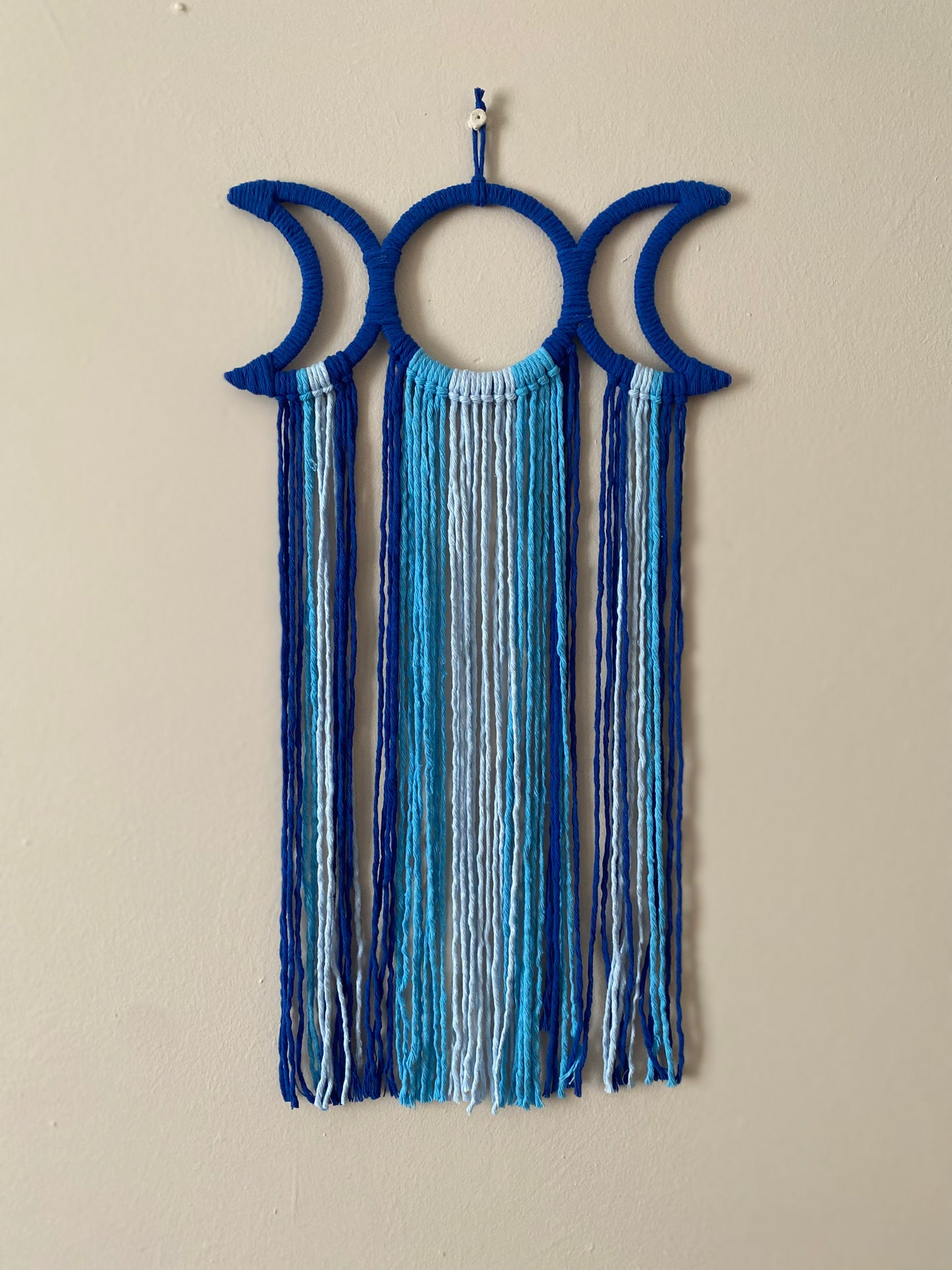 Handmade macramé wall hanging with triple moon goddess design, featuring recycled cotton cord in shades of blue, hanging against a beige wall.