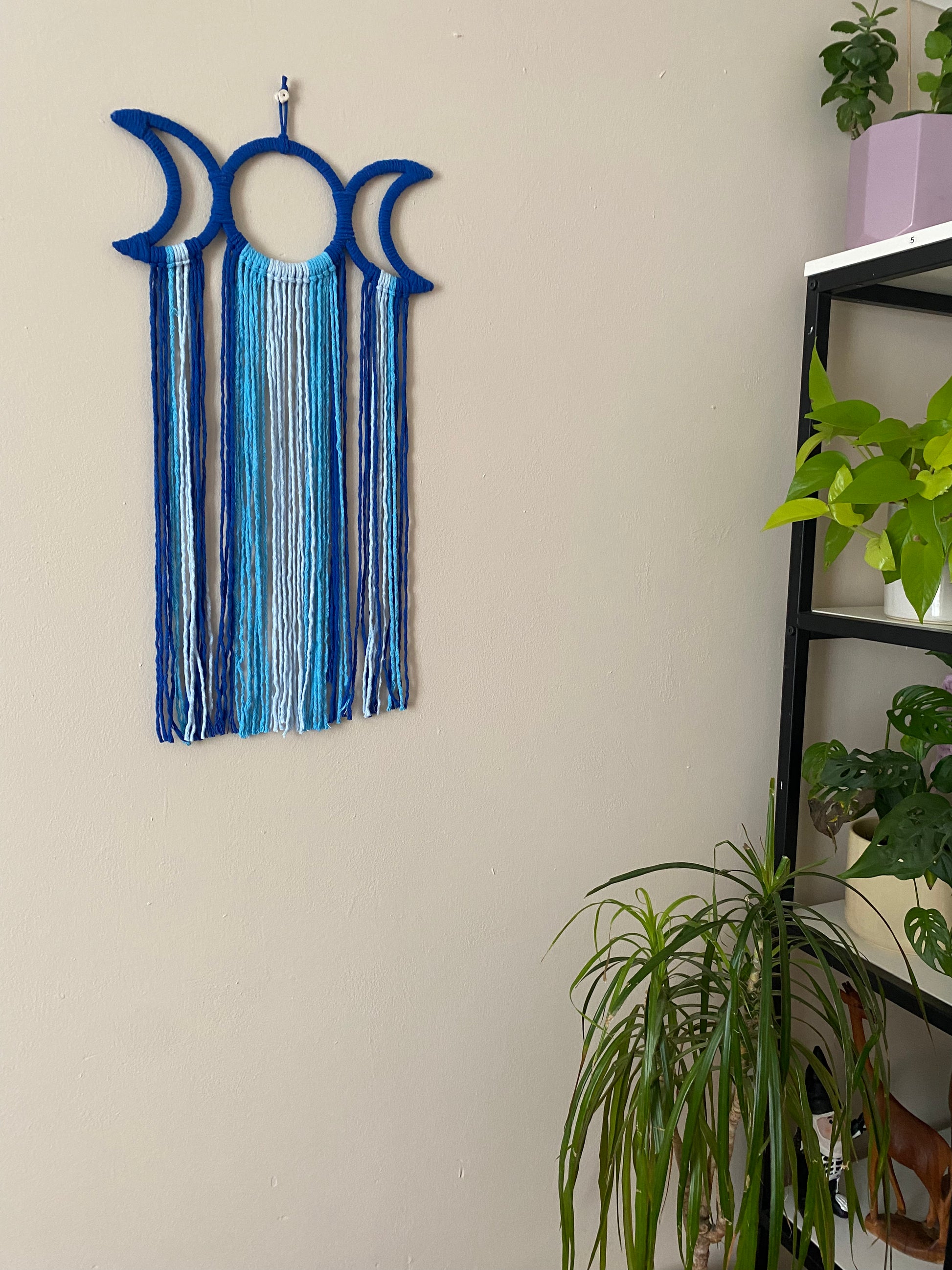 Handmade macramé wall hanging with triple moon goddess design, featuring recycled cotton cord in shades of blue, hanging against a beige wall, next to a tall shelving unit, which is full of houseplants.
