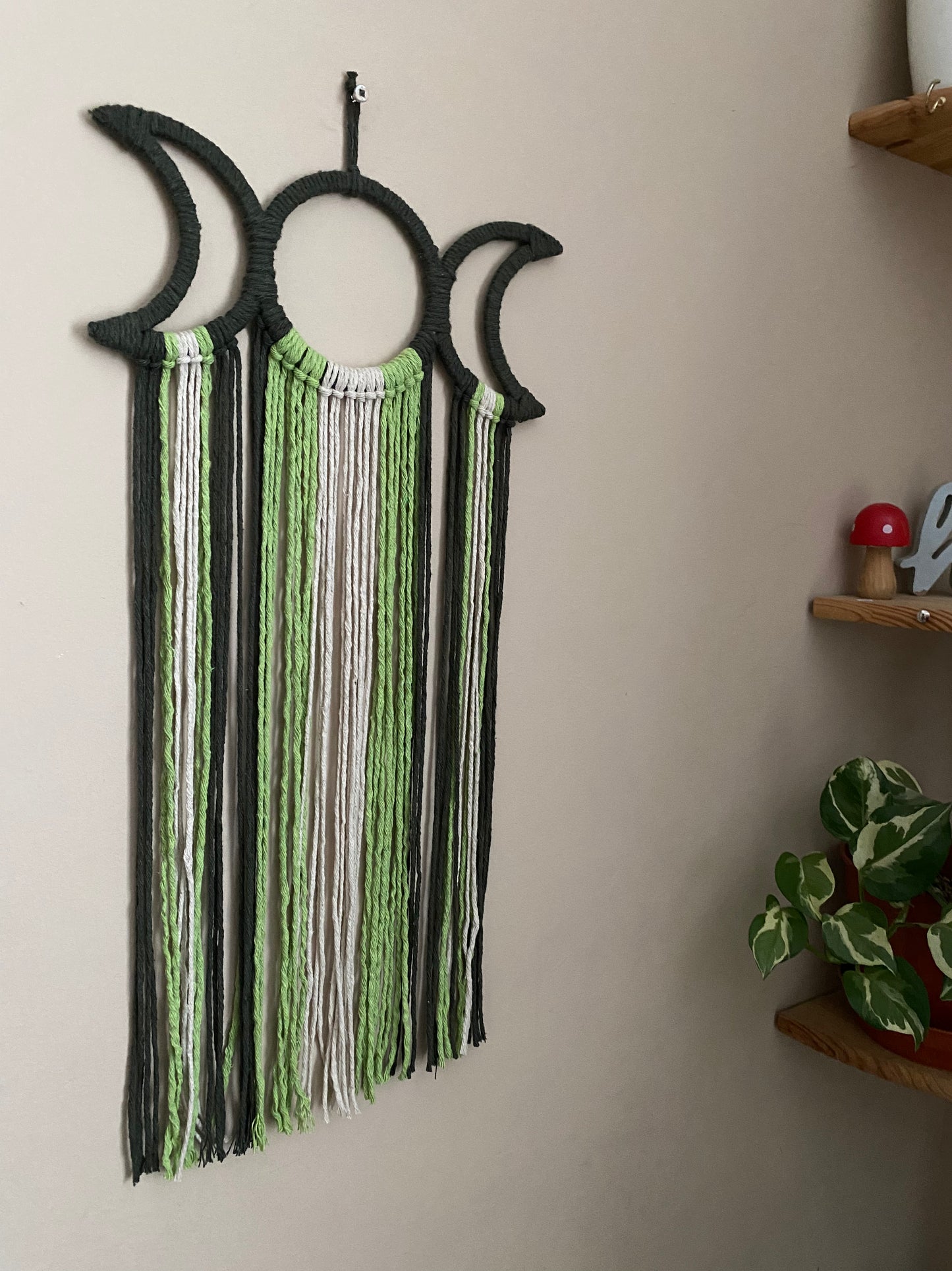 Handmade macramé wall hanging with triple moon goddess design, featuring recycled cotton cord in shades of green, hanging against a beige wall.