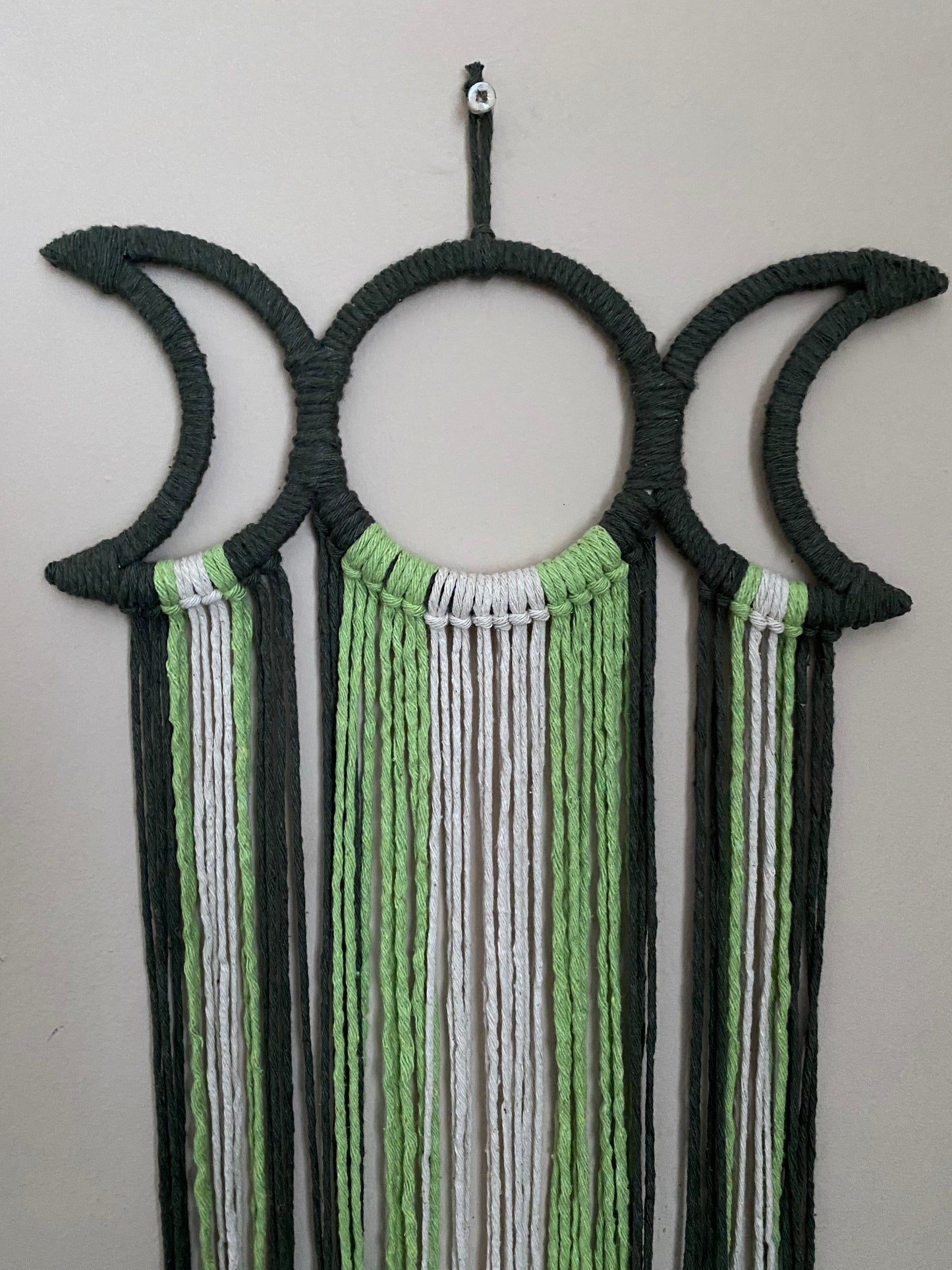 Close up of a handmade macramé wall hanging with triple moon goddess design, featuring recycled cotton cord in shades of green, hanging against a beige wall.