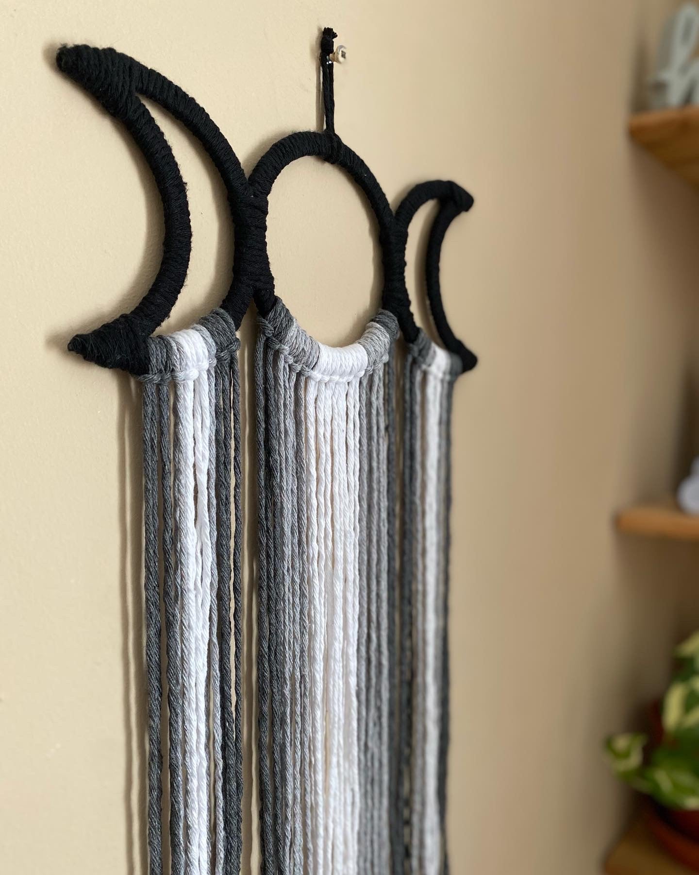 Close up of a handmade macramé wall hanging with triple moon goddess design, featuring recycled cotton cord in monochrome shades, hanging against a beige wall.