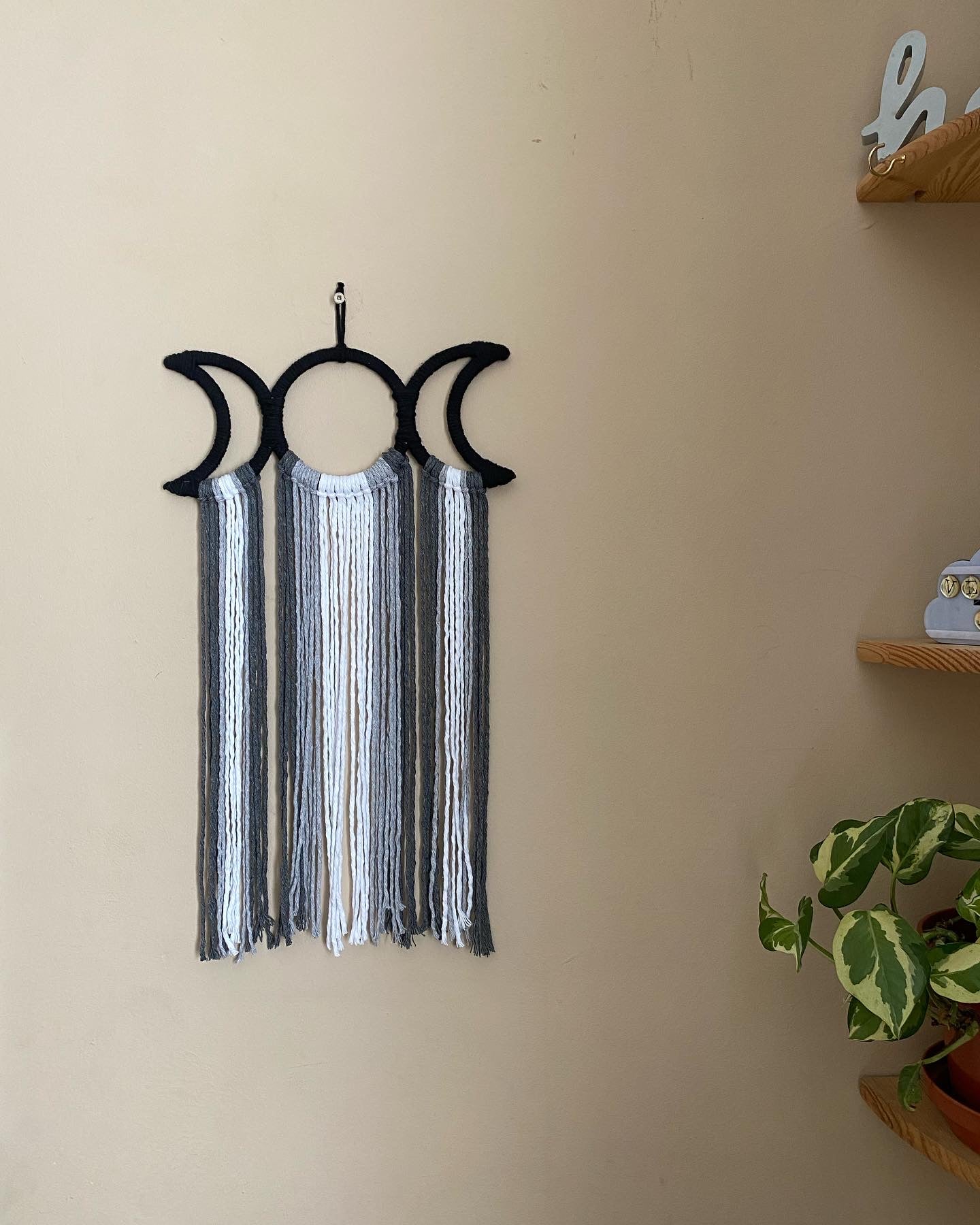 Handmade macramé wall hanging with triple moon goddess design, featuring recycled cotton cord in monochrome shades, hanging against a beige wall.