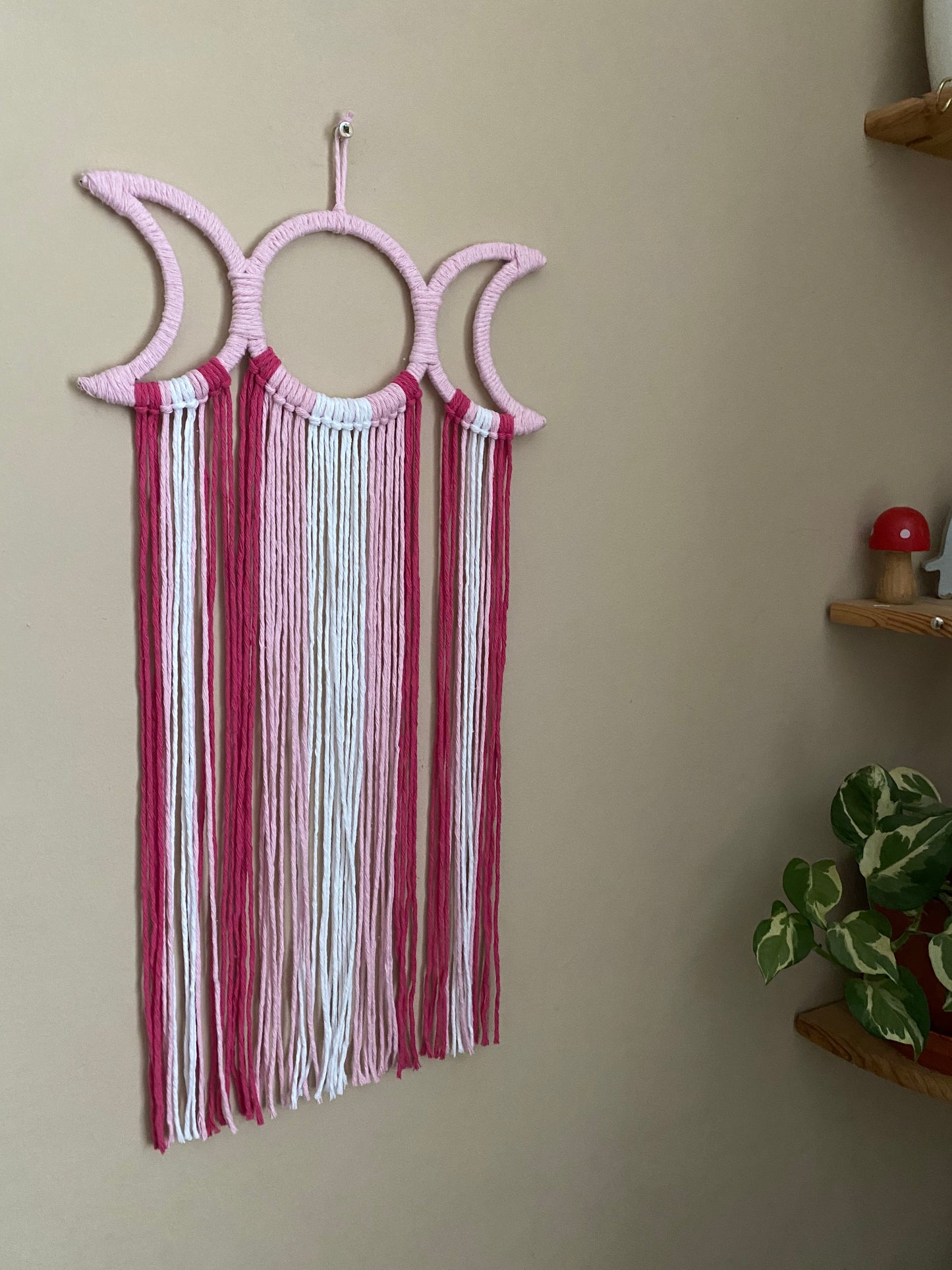 Handmade macramé wall hanging with triple moon goddess design, featuring recycled cotton cord in shades of pink, hanging against a beige wall.