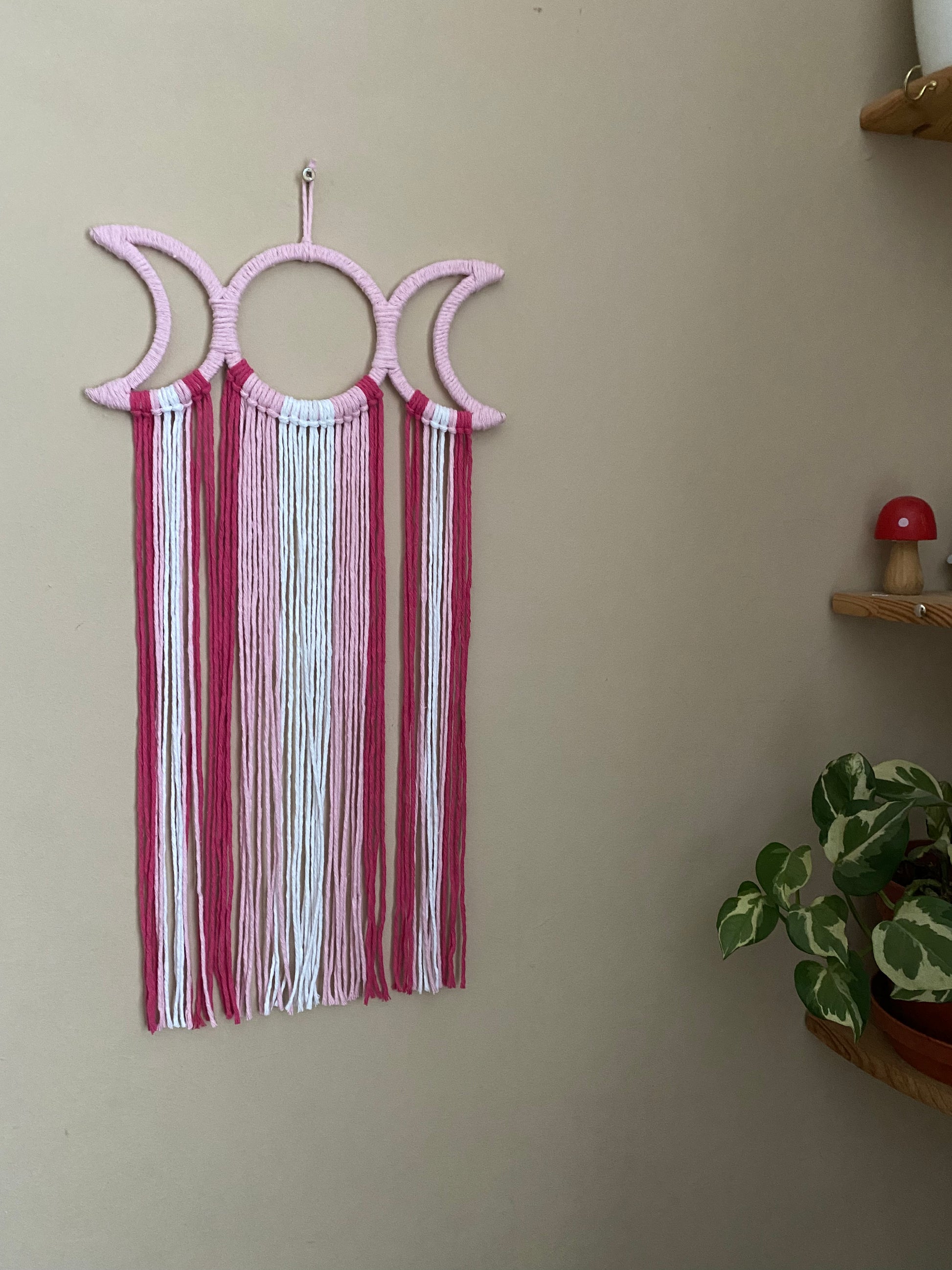 Handmade macramé wall hanging with triple moon goddess design, featuring recycled cotton cord in shades of pink, hanging against a beige wall.