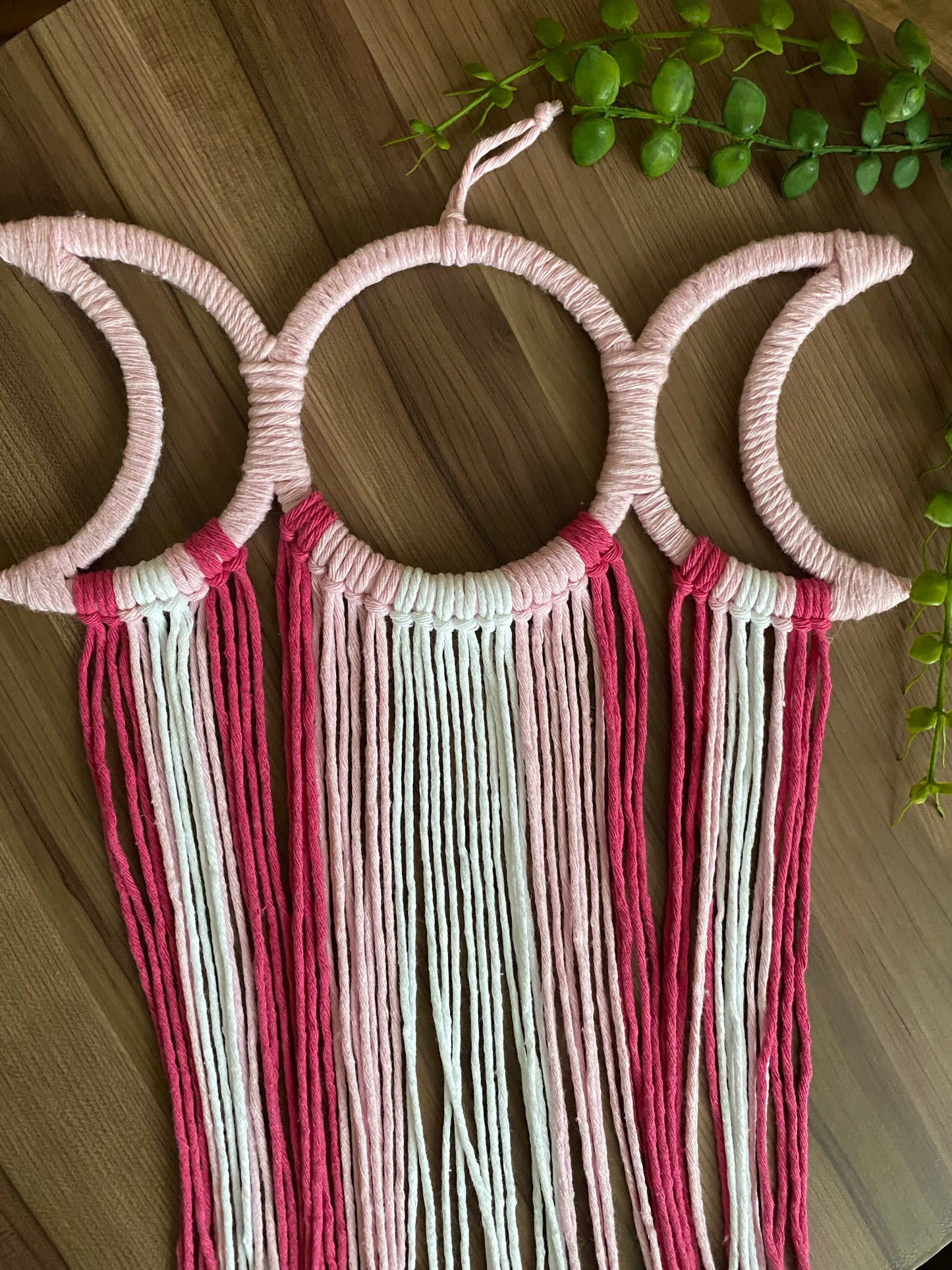 Handmade macramé wall hanging with triple moon goddess design, featuring recycled cotton cord in shades of pink, laid flat on a wooden surface with some green plant vines.
