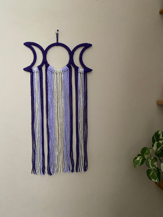 Handmade macramé wall hanging with triple moon goddess design, featuring recycled cotton cord in shades of purple, hanging against a beige wall.