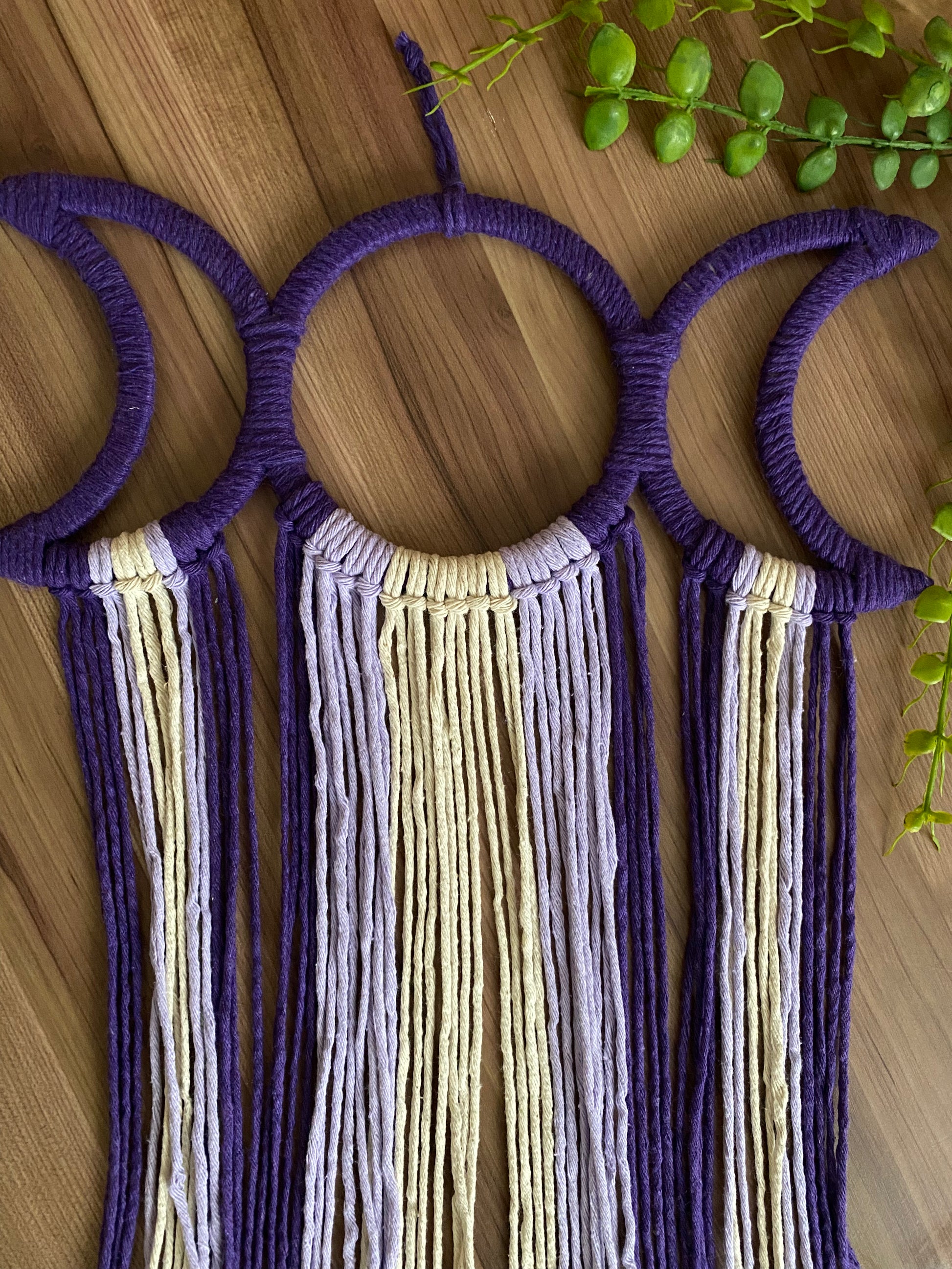 Handmade macramé wall hanging with triple moon goddess design, featuring recycled cotton cord in shades of purple, laid flat on a wooden surface with some green plant vines.
