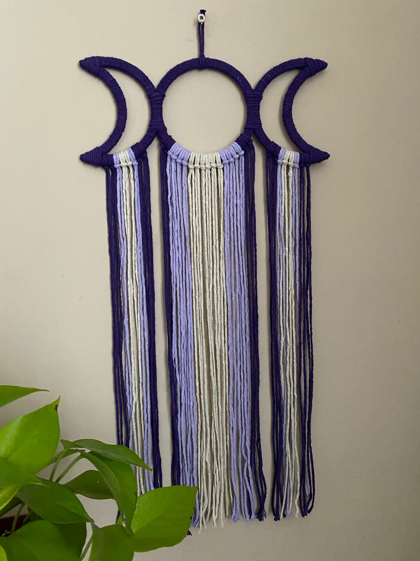 Handmade macramé wall hanging with triple moon goddess design, featuring recycled cotton cord in shades of purple, hanging against a beige wall. There is a bright green plant visible in the bottom left corner.
